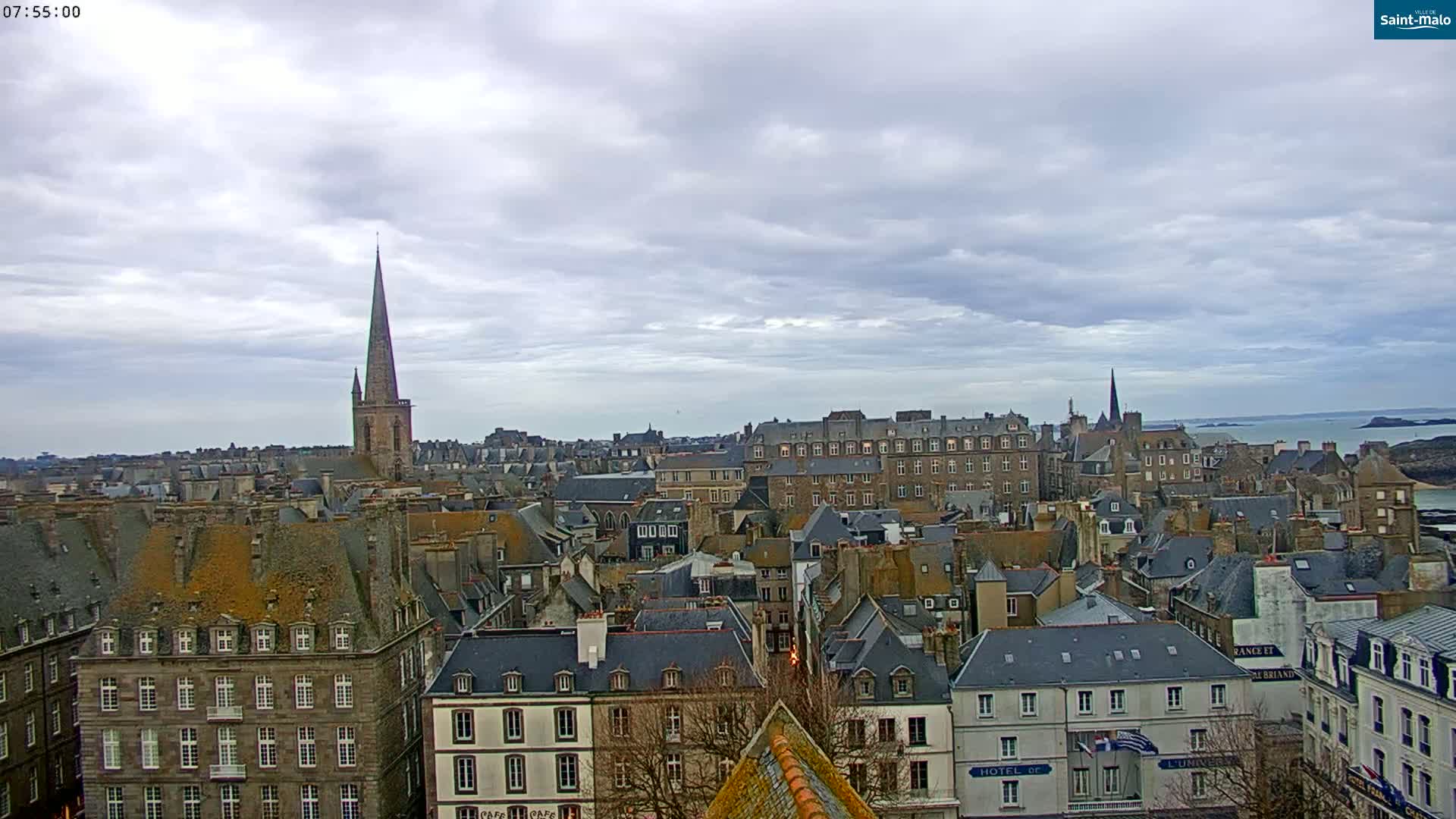 A cloudy day overlooks a coastal city with numerous stone buildings, a prominent church spire, and a view of the ocean in the distance.