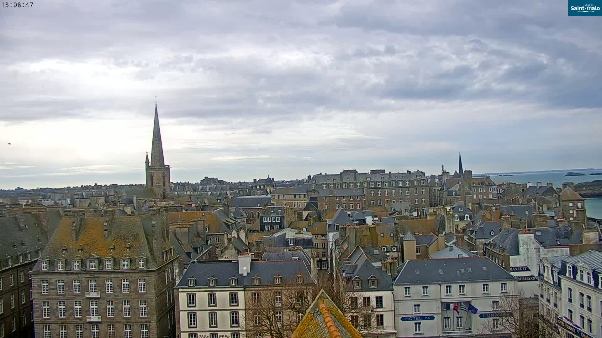 An overcast sky hangs over a cityscape of closely packed buildings with gray slate roofs, punctuated by a tall church spire and a body of water in the distance.