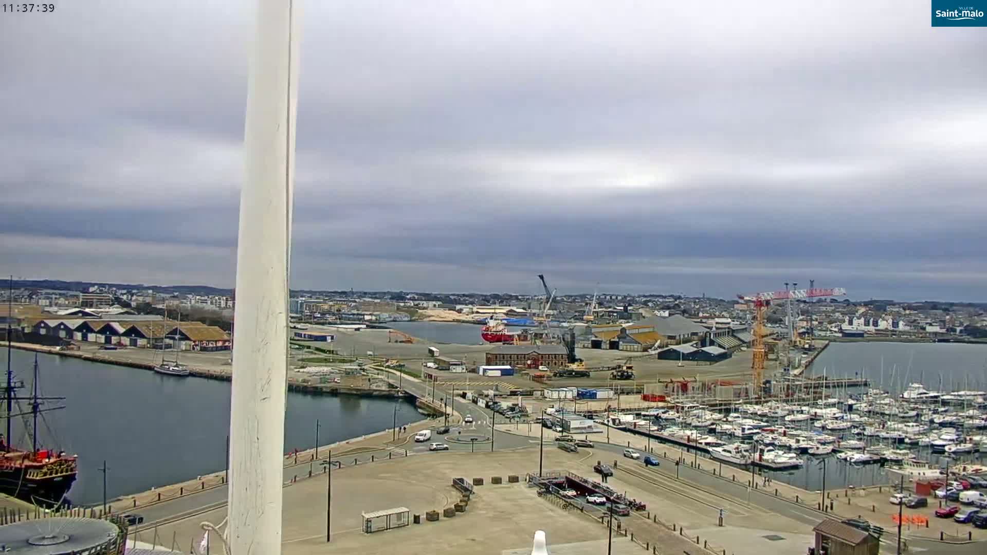 A high-angle view of a harbor on an overcast day, showing a marina filled with sailboats, a shipyard with cranes, and a large ship docked nearby.