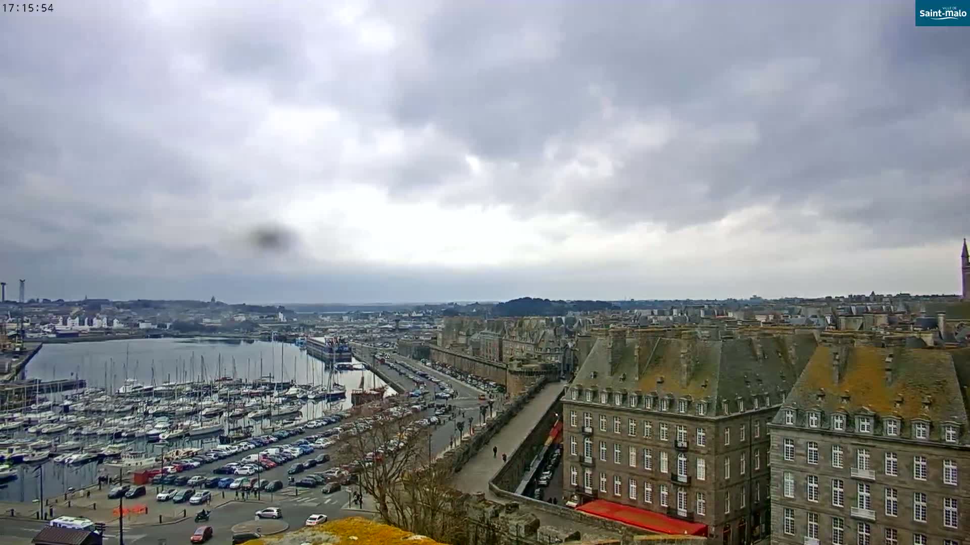 A harbor filled with sailboats is visible from an elevated vantage point overlooking a city with overcast skies.