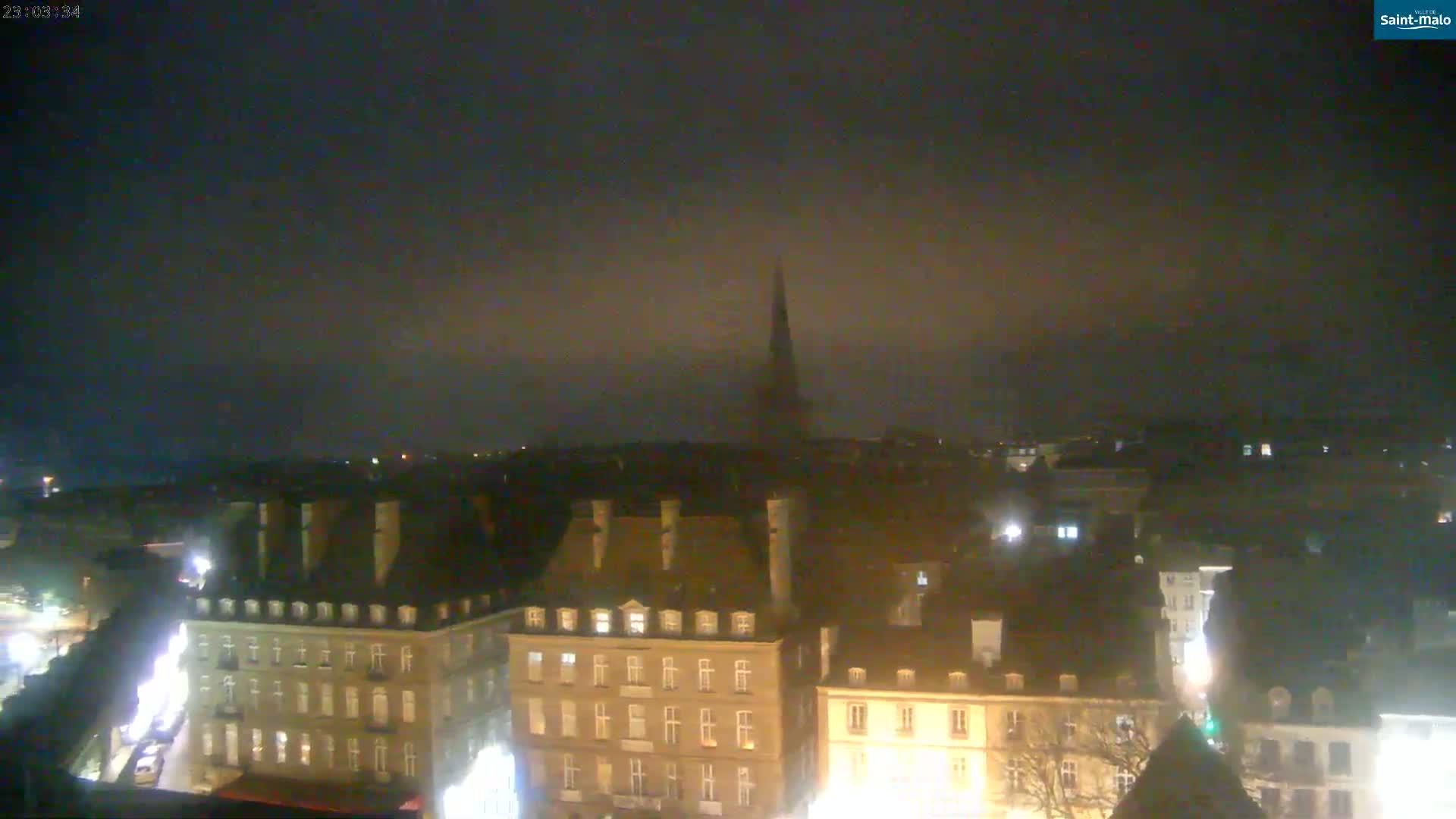 A nighttime view of a city skyline, shrouded in a hazy fog, shows a collection of buildings with illuminated windows, and a tall, pointed spire in the distance.