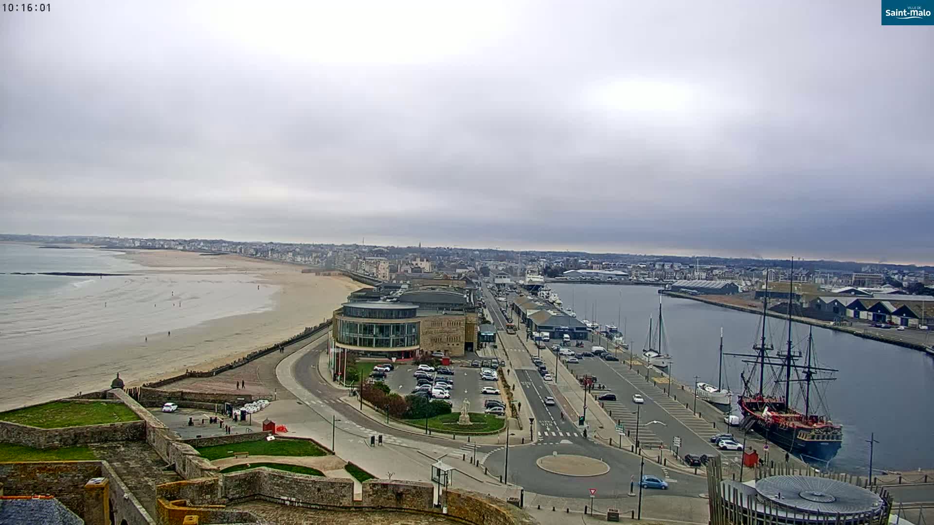 A coastal city with a long sandy beach, a harbor filled with boats, and a large sailing ship is viewed from a high vantage point on an overcast day.