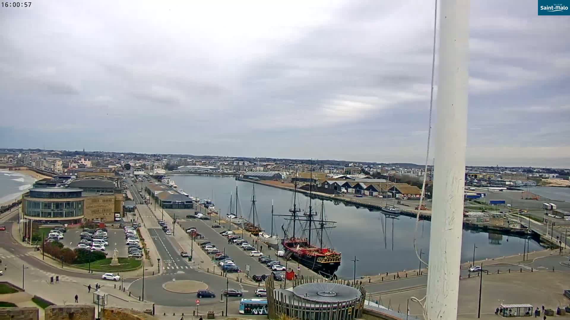 A harbor with a large sailing ship, many parked cars, and buildings is visible under an overcast sky.