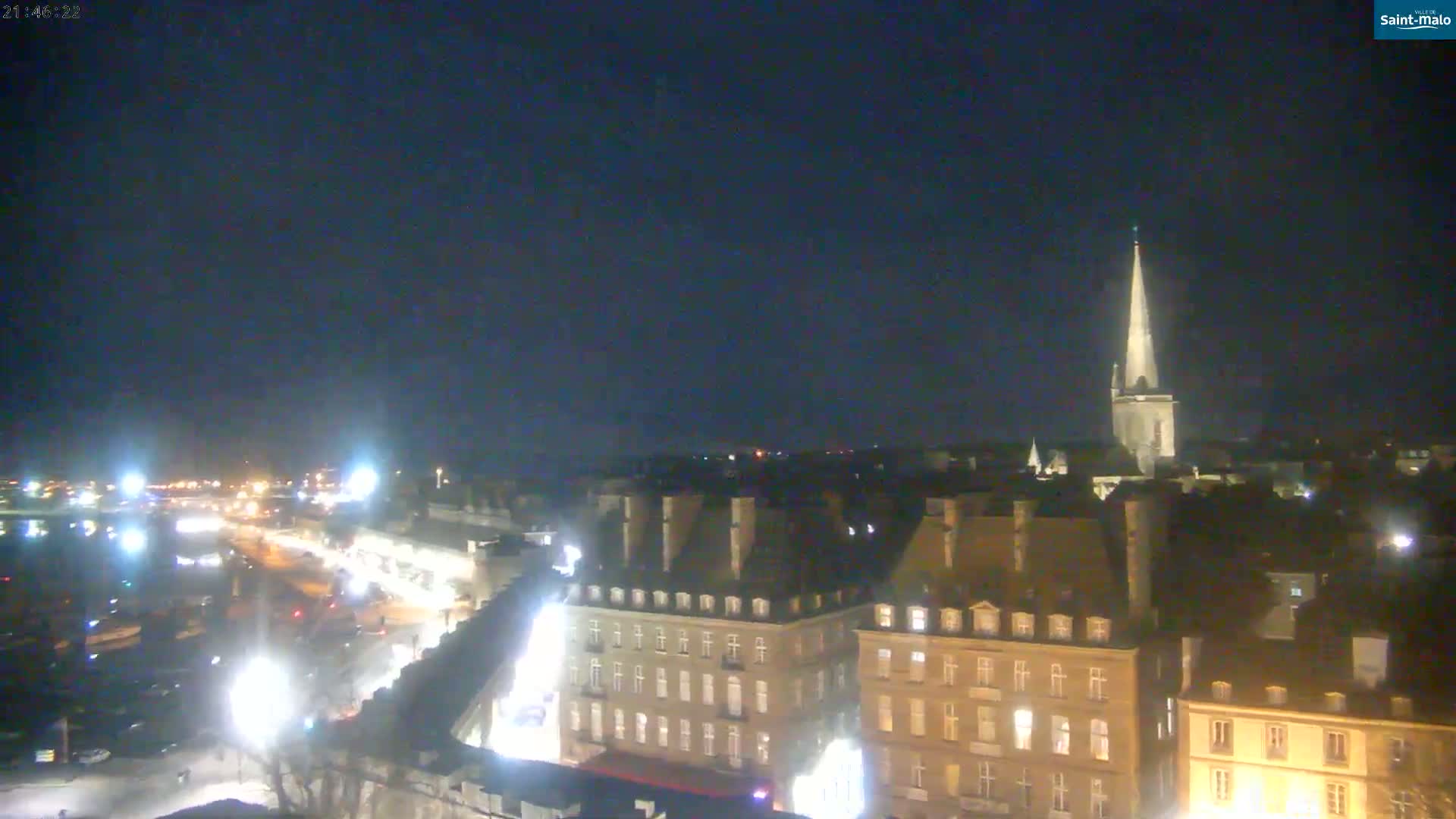 A nighttime, aerial view of a city with a tall church spire in the distance, under a clear, dark sky.