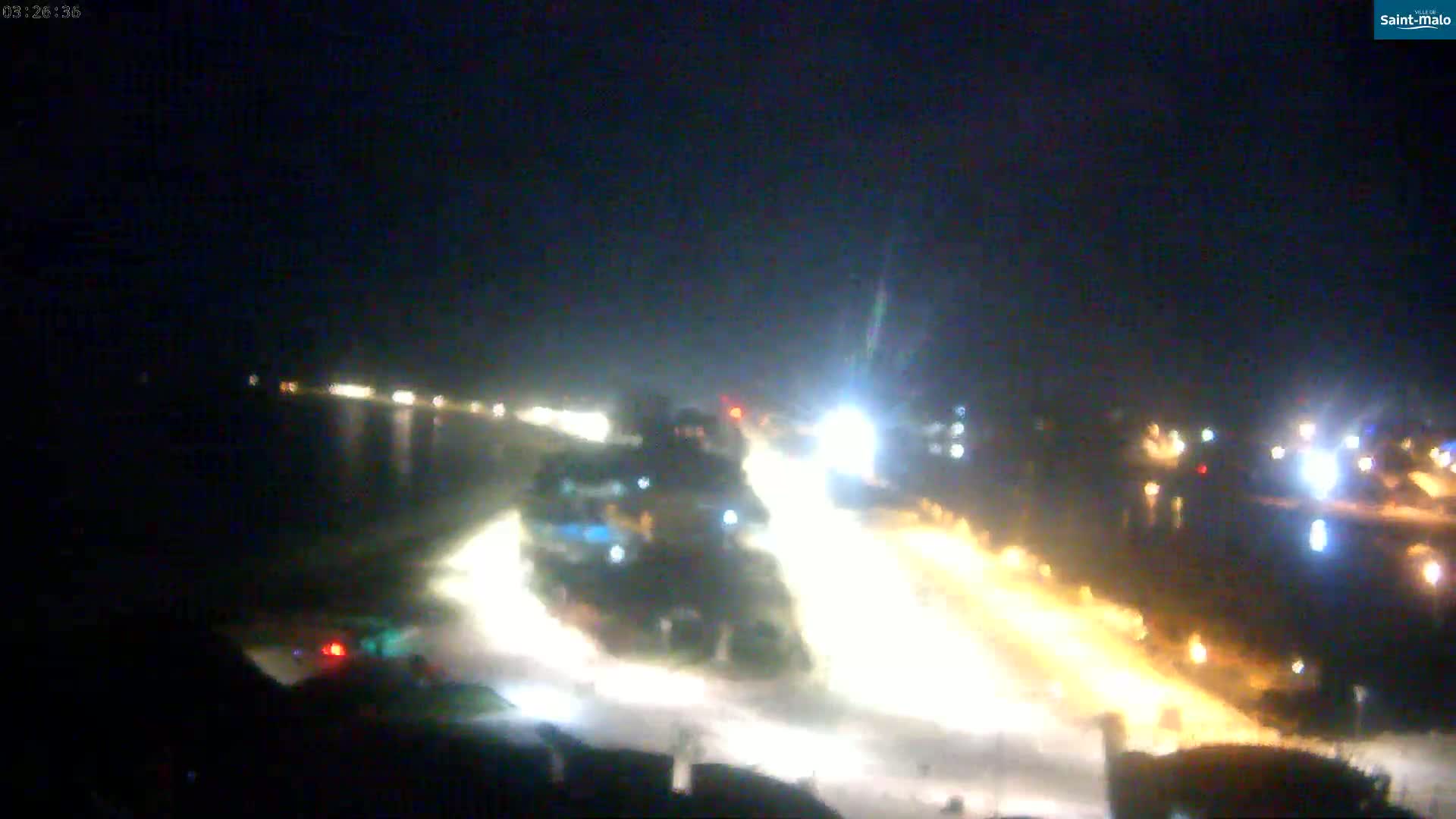 A nighttime, blurry long shot shows a roadway with bright lights illuminating the area near a body of water.