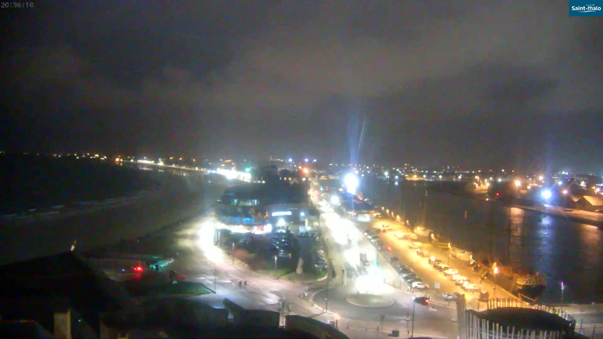 A nighttime aerial view of a coastal town shows a beach, roads with cars, and buildings illuminated under a dark, cloudy sky.