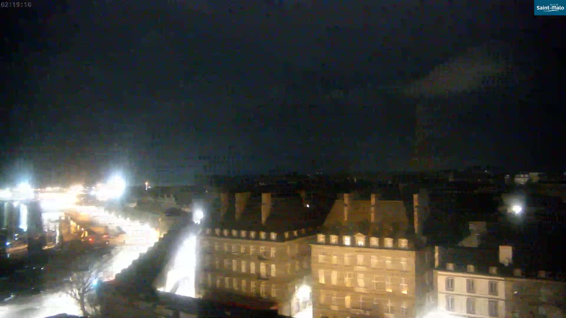 A nighttime aerial view of a city with dimly lit buildings under a dark, cloudy sky.
