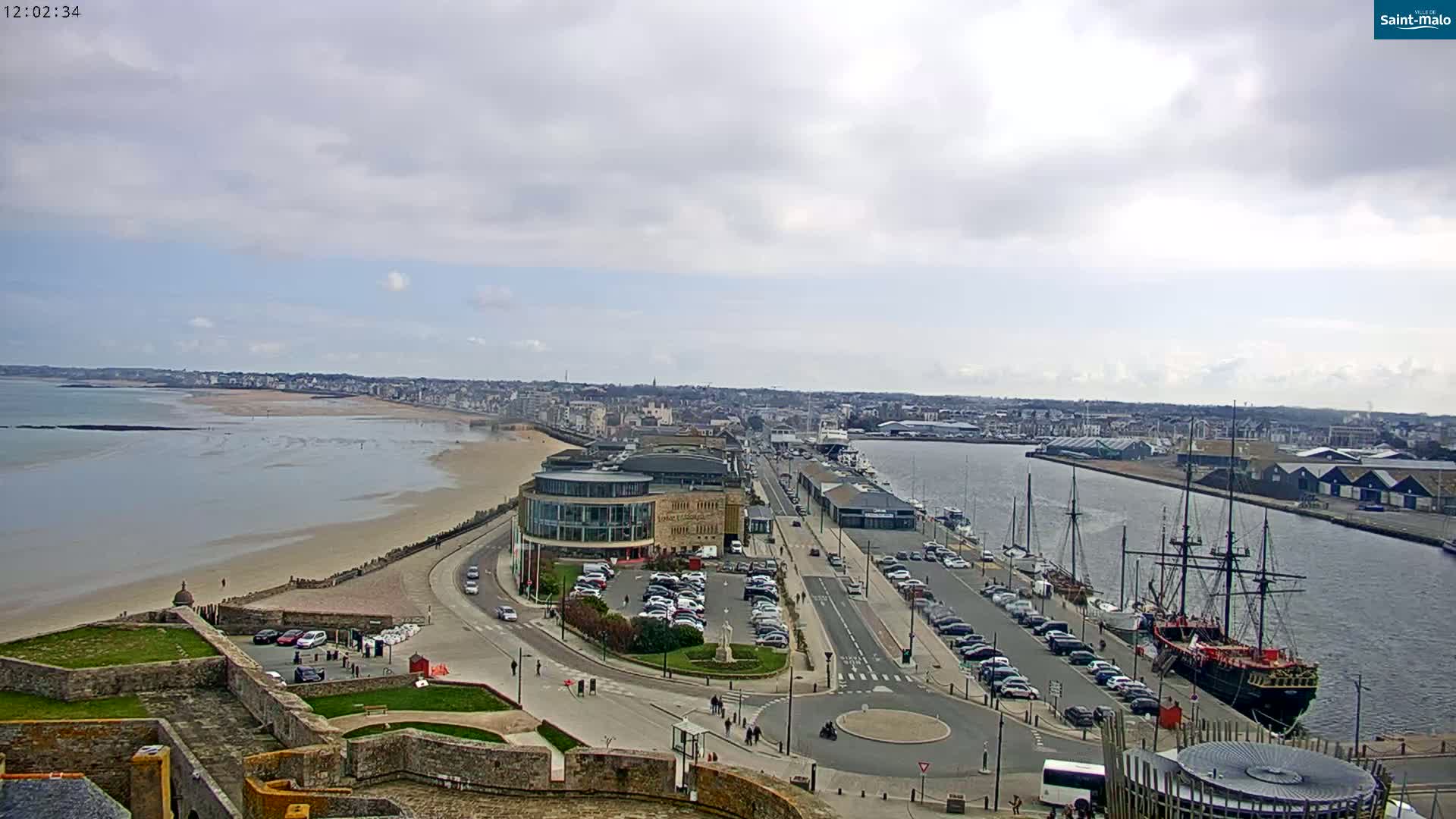 A coastal city with a harbor filled with ships is seen on an overcast day, from a high vantage point overlooking a sandy beach and stone fortifications.