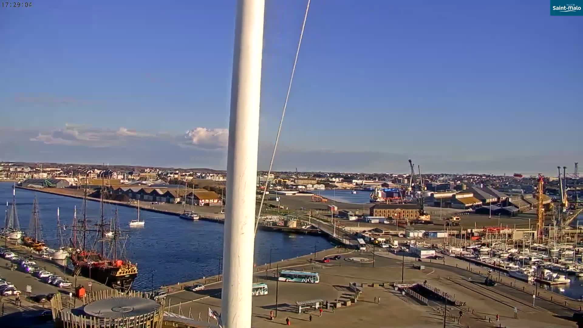 A mostly sunny harbor scene shows a large sailing ship docked next to several smaller vessels, with a city and numerous industrial buildings stretching into the distance.