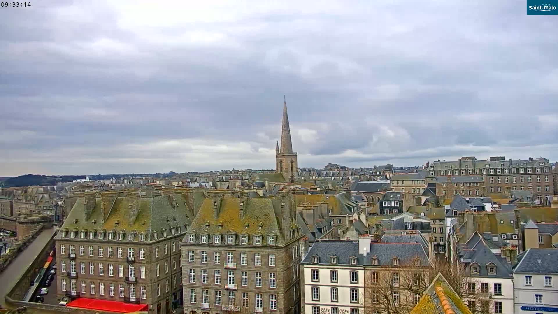 A sprawling cityscape of grey-roofed buildings under a cloudy sky, featuring a prominent church spire in the mid-ground.