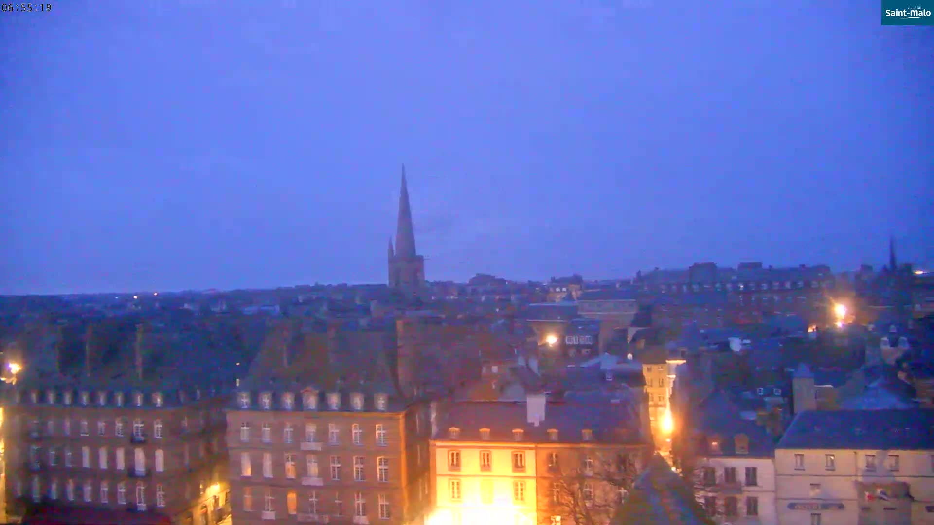 A twilight view of a city skyline with numerous buildings and a tall church steeple, under a clear, dusky sky.