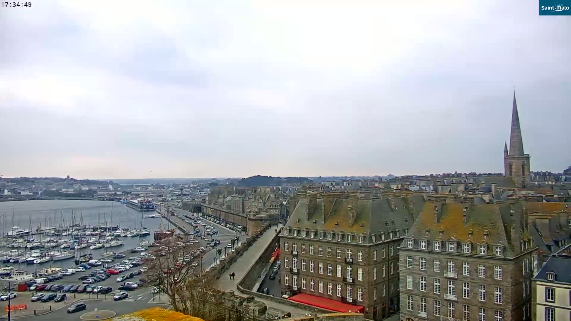 A harbor filled with sailboats is visible beneath a cloudy sky, with a city of stone buildings and a tall church spire in the background.