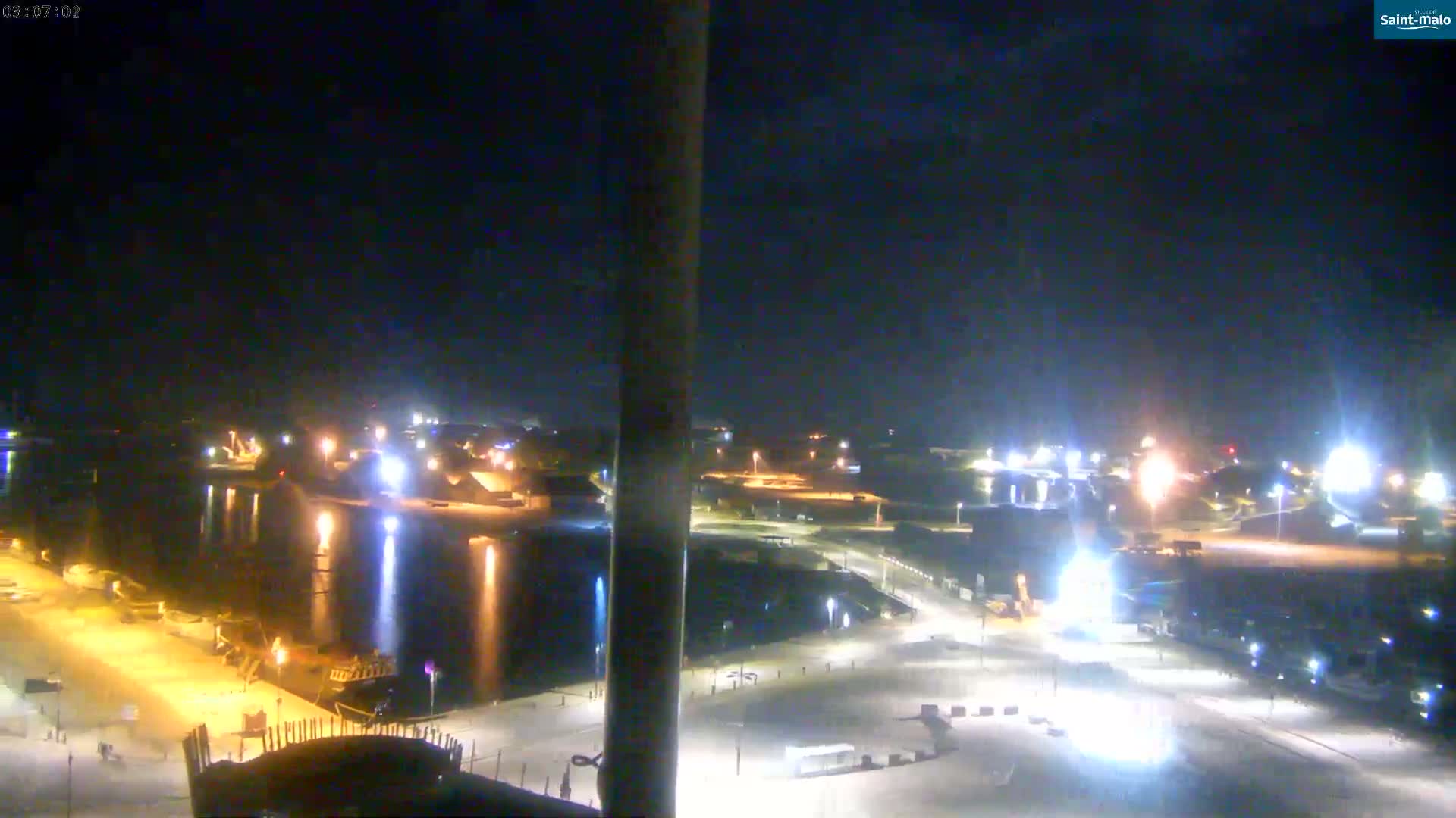 A nighttime, brightly lit harbor scene with calm water reflecting the lights, under a dark sky.