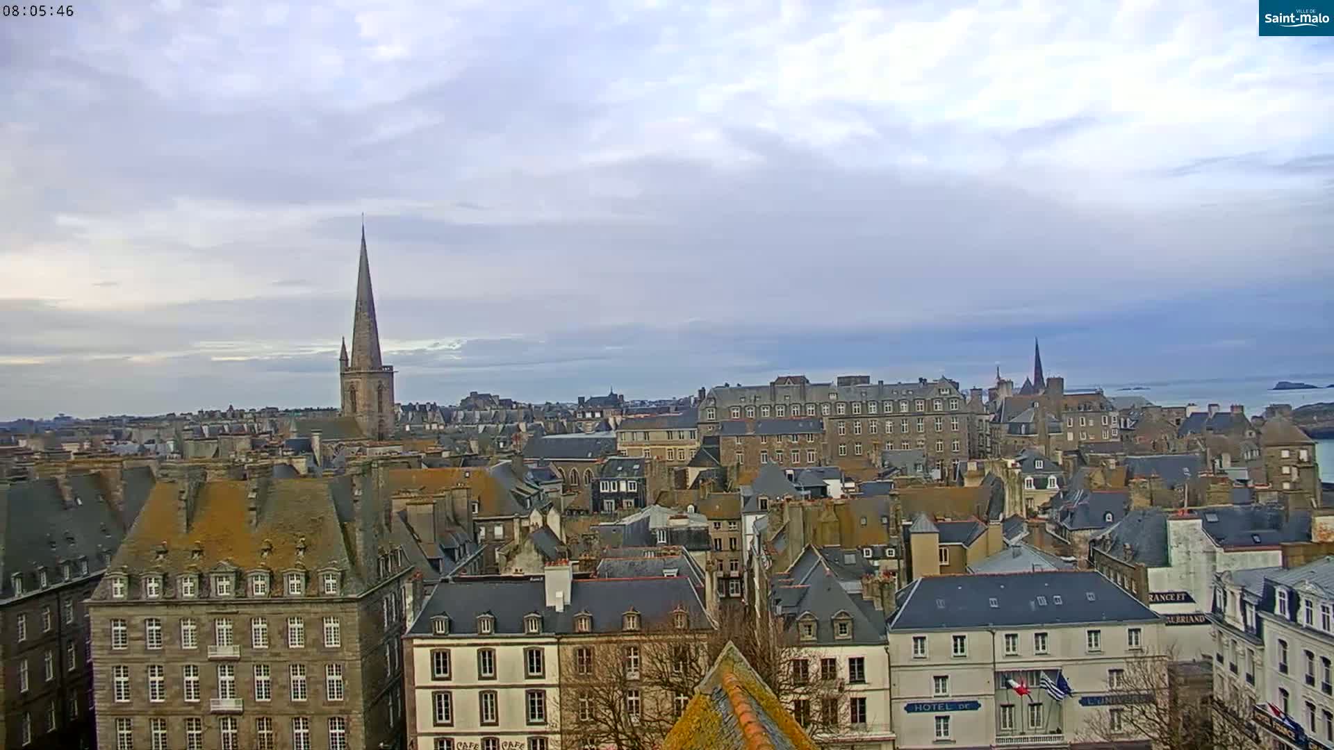 A panoramic view of a European city with many grey-roofed buildings under a cloudy sky, featuring a prominent church spire.