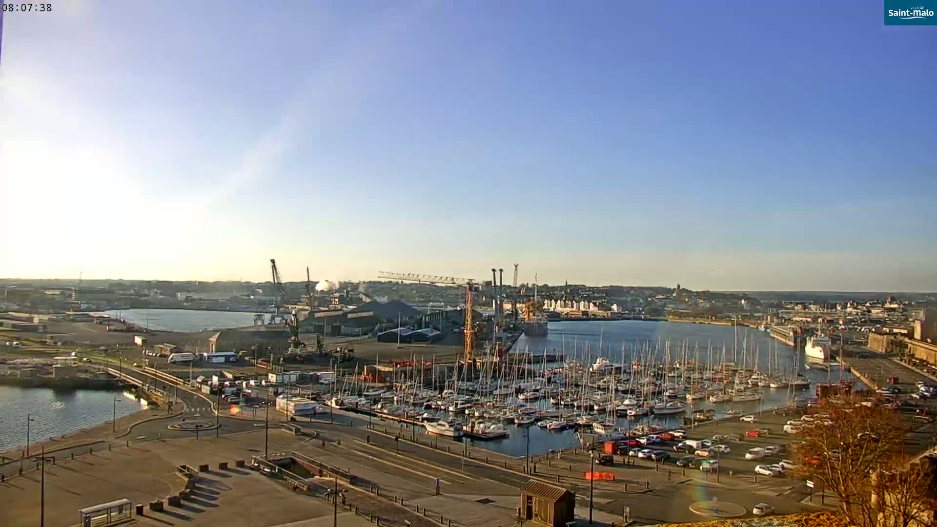 A harbor filled with numerous sailboats and yachts under a clear, sunny sky, with cranes and industrial buildings visible in the background.
