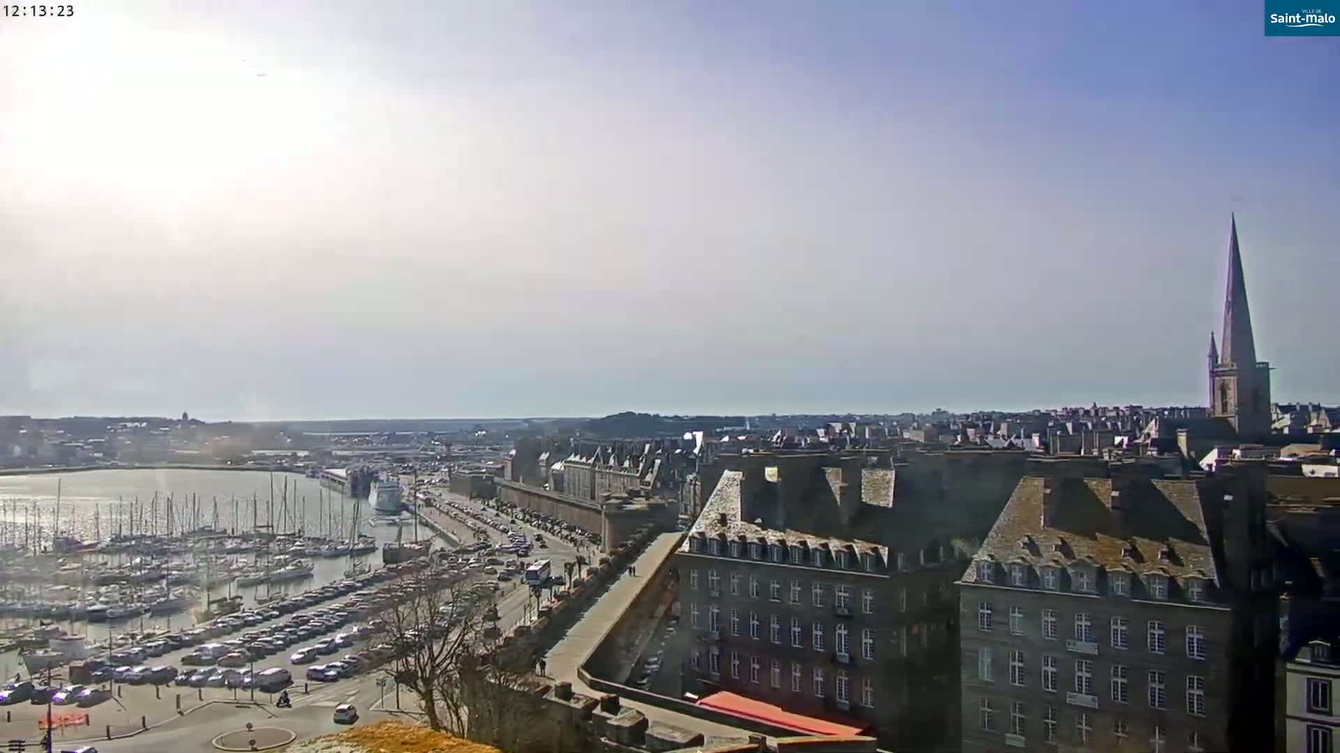 A sunny day reveals a harbor filled with boats, a city with a tall church spire, and a large parking lot, all viewed from a high vantage point.
