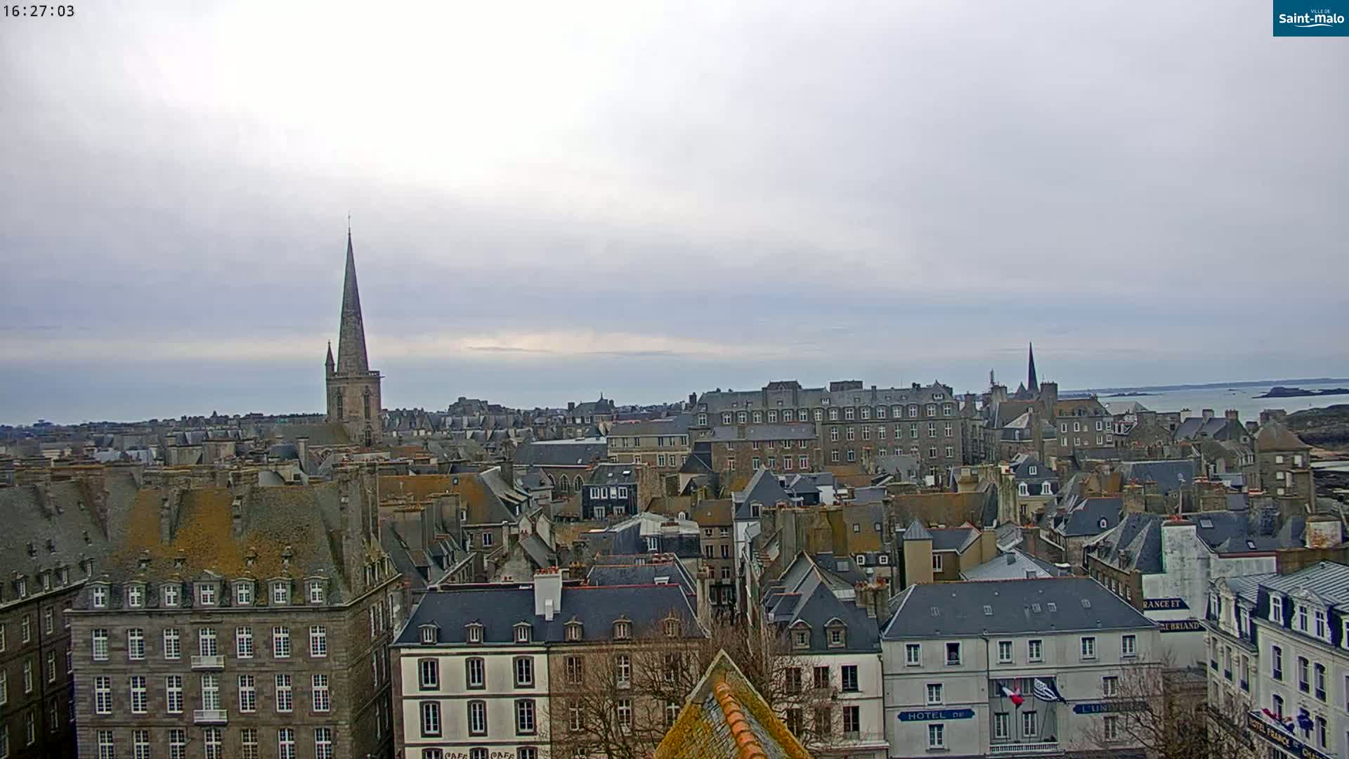 A panoramic view of a city with many grey-roofed buildings under an overcast sky, showing a coastline and body of water in the distance.