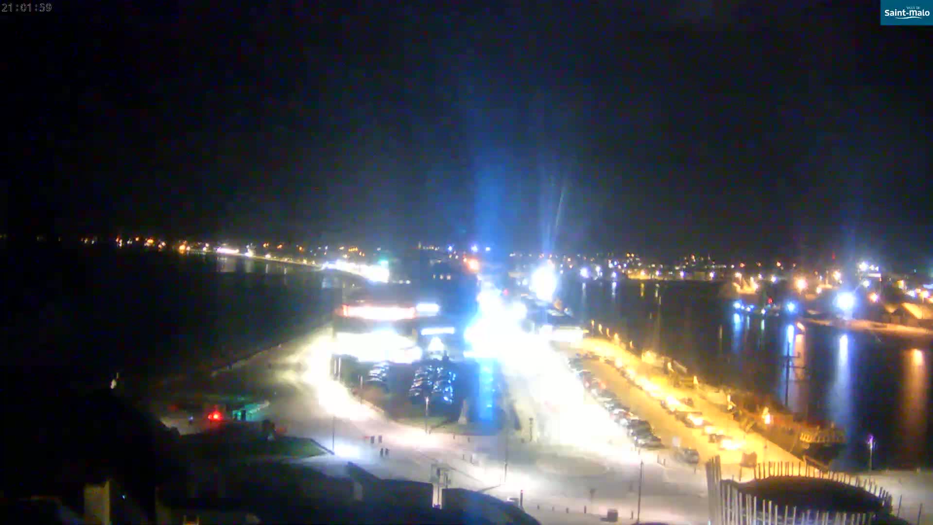 A nighttime, brightly lit waterfront scene shows a body of water reflecting city lights, with a parking lot and buildings visible on land.