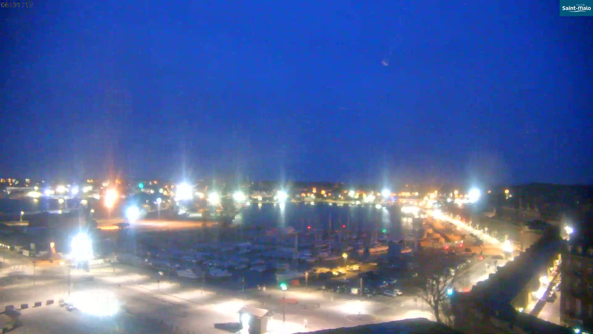 A nighttime, harbor-side view of a town with many boats in the water and lights illuminating the streets and docks under a clear, dark blue sky.