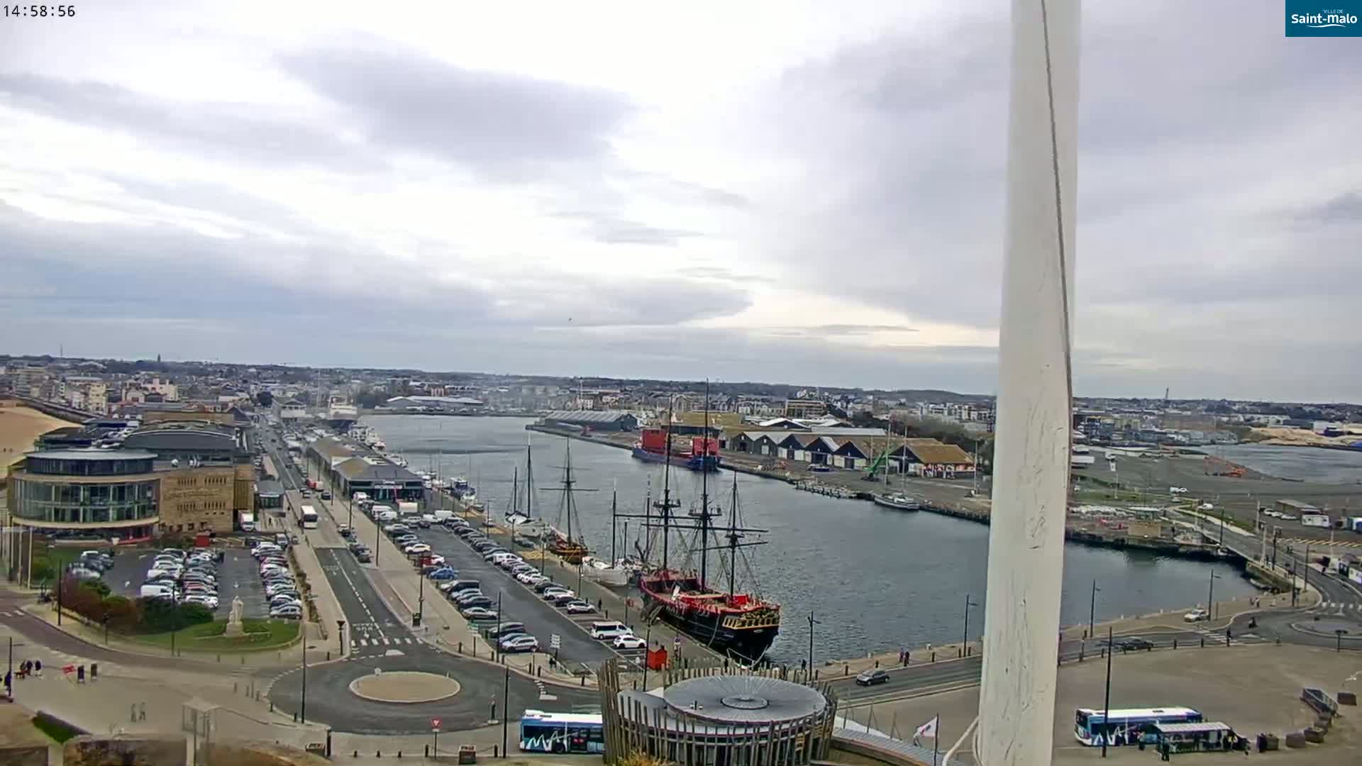 An overcast day reveals a harbor scene with a large sailing ship docked near a city, alongside parked cars and buildings.