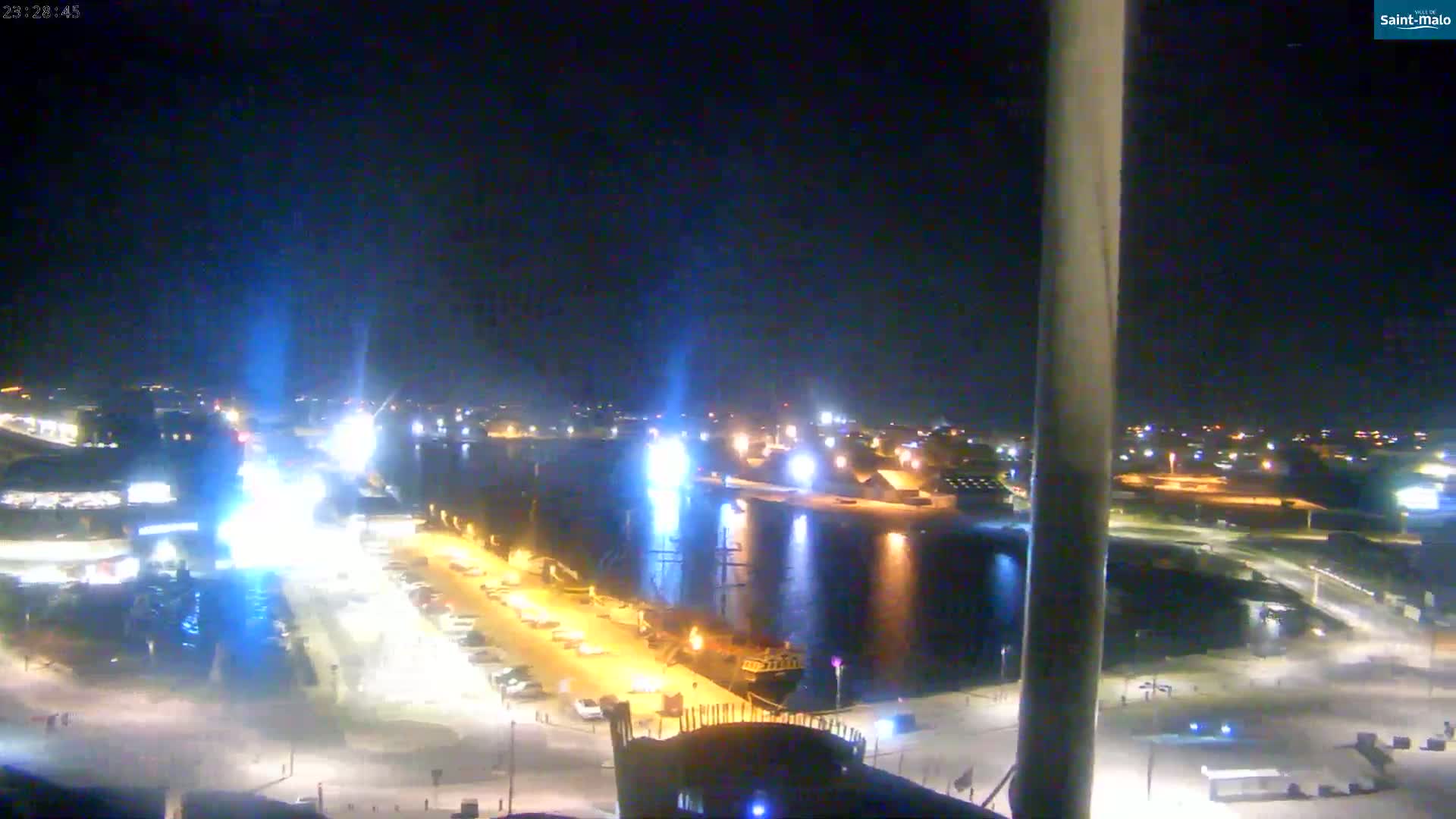 A nighttime view of a harbor with boats docked, illuminated buildings, and parking lots, under a clear sky.