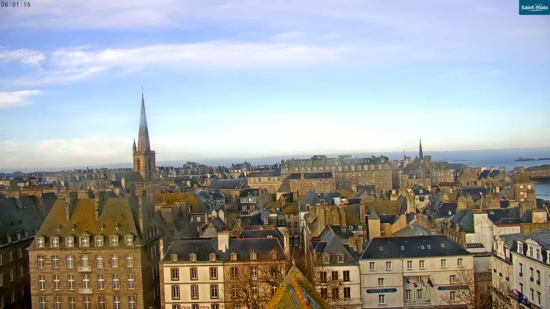 A panoramic view of a coastal city on a partly cloudy day, showing many stone buildings with steeply pitched roofs, and a tall church steeple in the center.