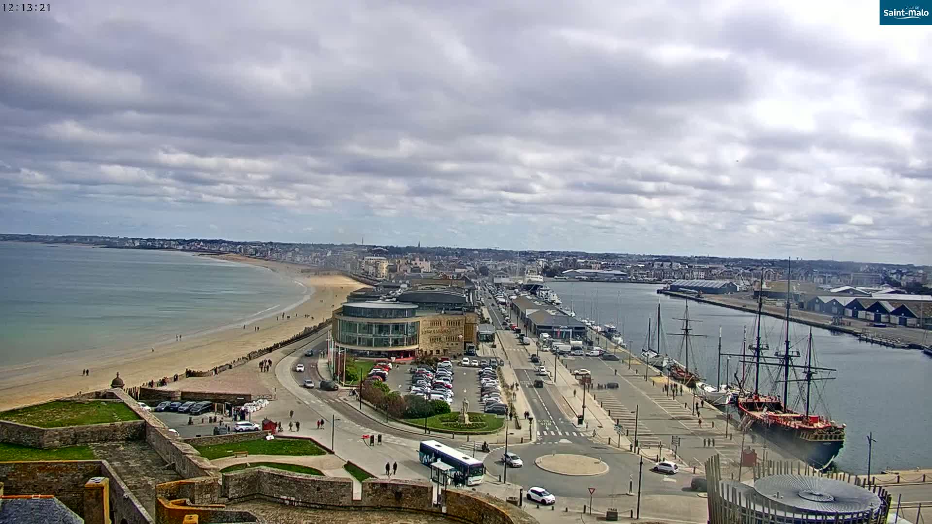 An aerial view shows a coastal city with a sandy beach, harbor filled with boats, and a large sailing ship docked, under a mostly cloudy sky.