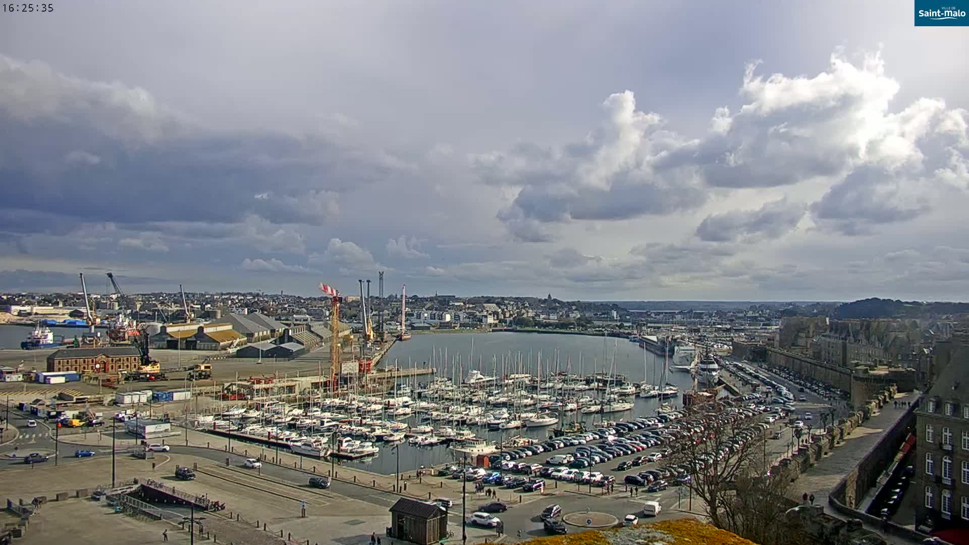 A harbor filled with numerous sailboats and cars parked along a waterfront, under a partly cloudy sky.