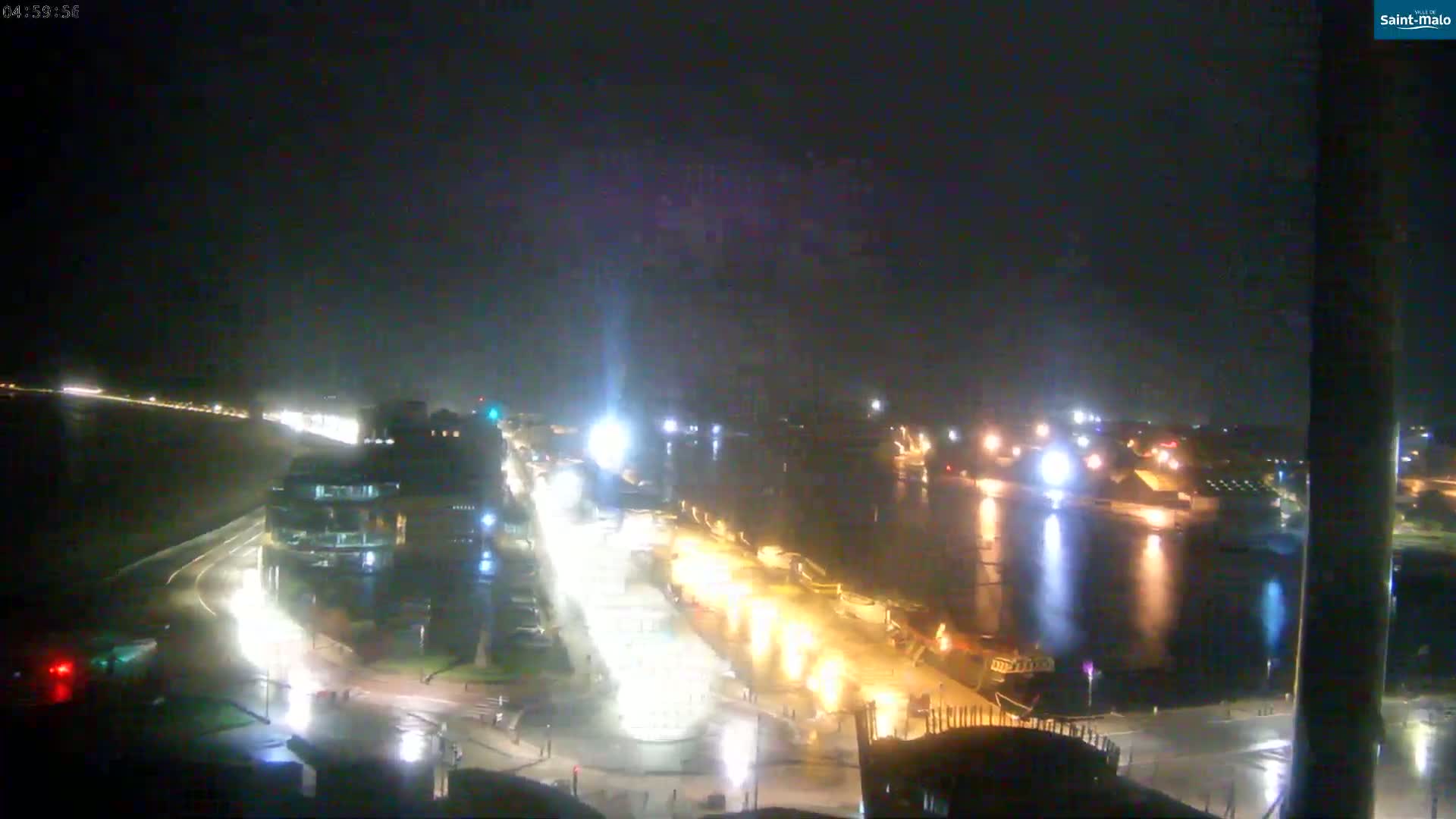 A nighttime, wet view of a city's waterfront at night, showing brightly lit streets and buildings alongside a dark body of water with lights reflecting on it.