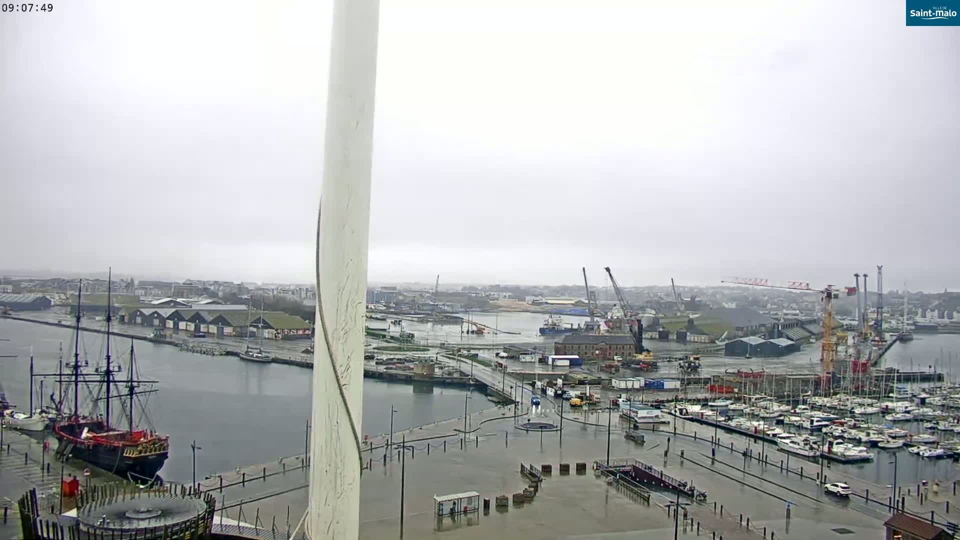 A rainy overcast day reveals a harbor scene with a large sailing ship, numerous smaller boats docked at a marina, several cranes, and industrial buildings.