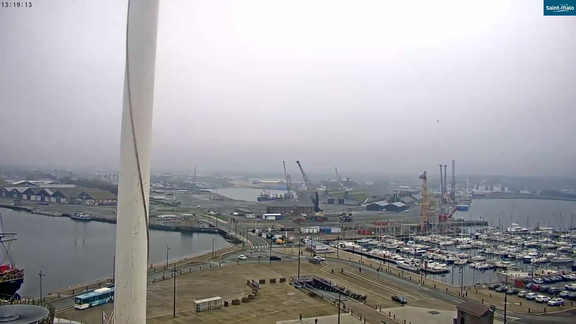 A hazy view shows a harbor with many boats docked, industrial areas, and a large, tall white structure in the foreground.