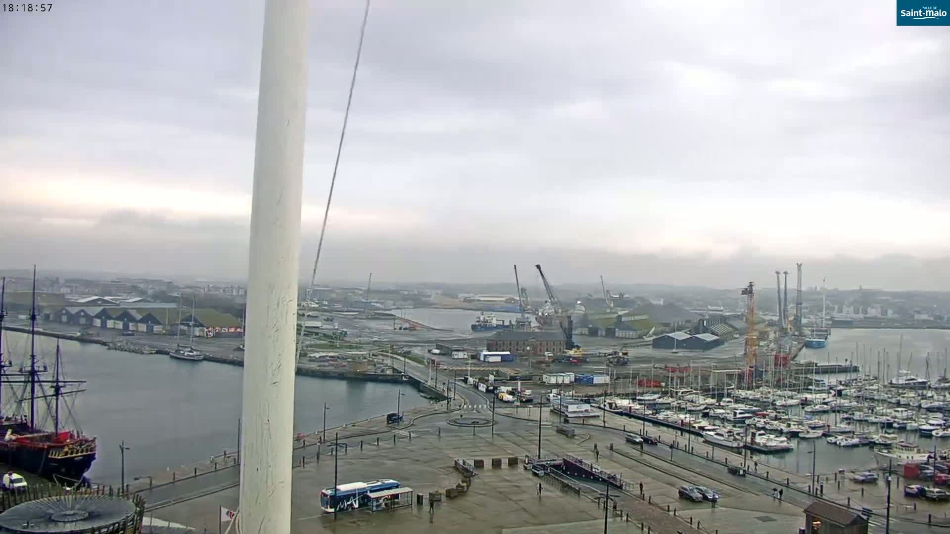 A harbor scene on an overcast day shows a large body of water with many docked boats and ships, some cranes, and industrial buildings, all viewed from an elevated position near a tall, white structure.