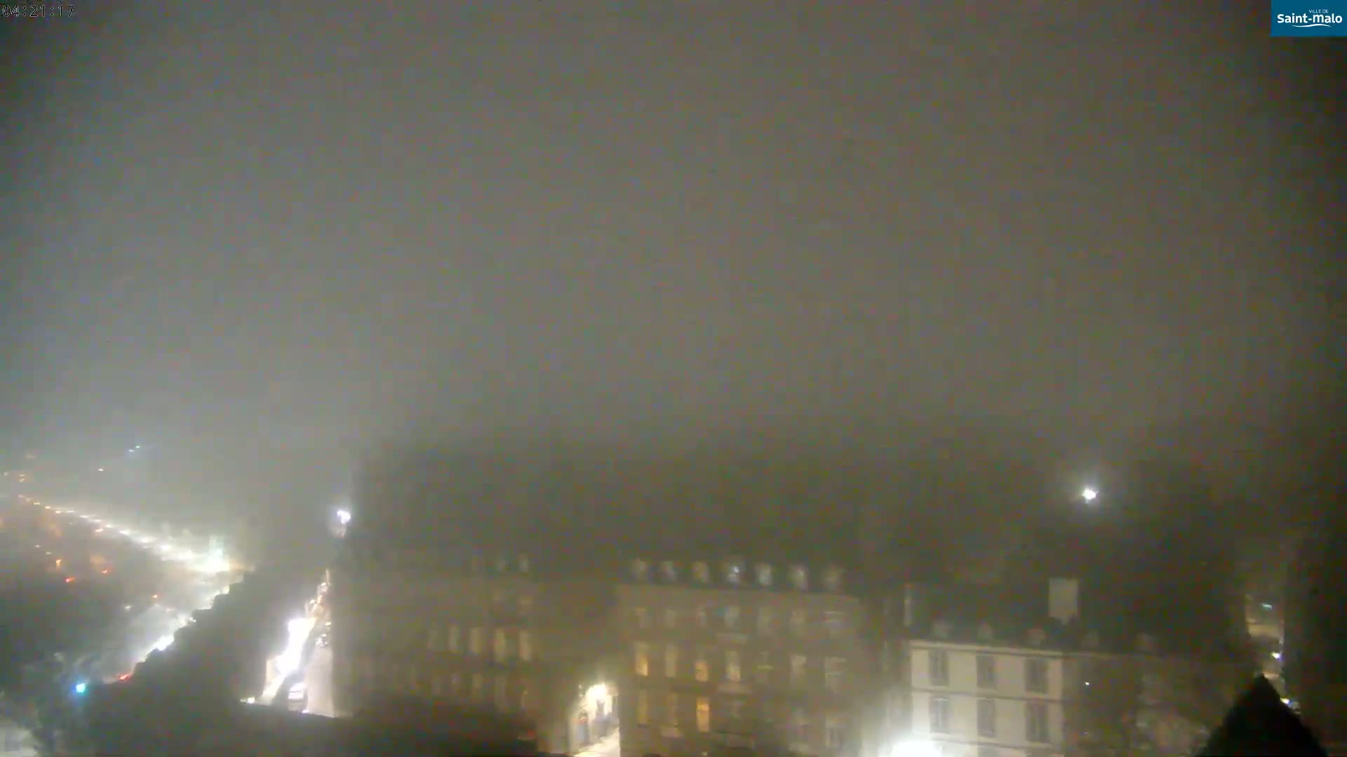 A nighttime, foggy view of a city street with buildings faintly visible through the heavy fog.