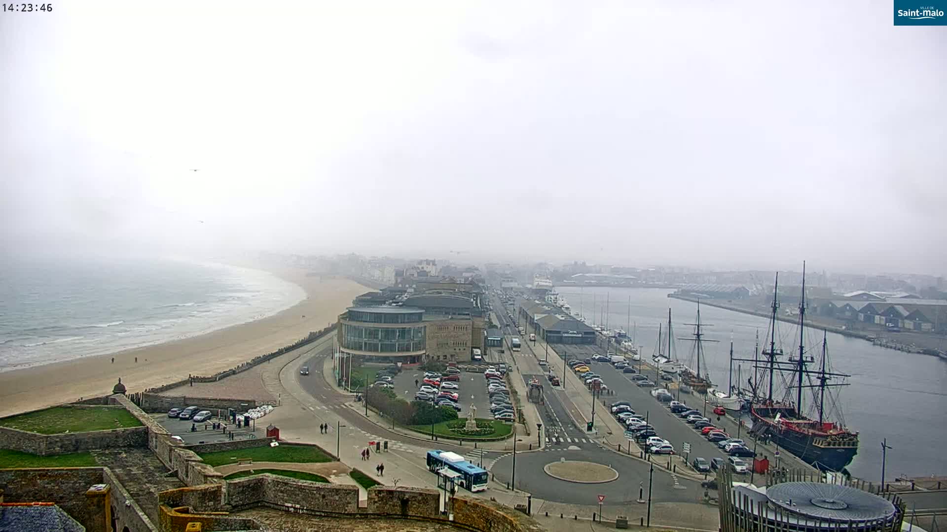 A coastal city scene on a foggy day shows a sandy beach, a harbor with several ships, including a large, dark-hulled sailing vessel, and a parking lot near a large building.