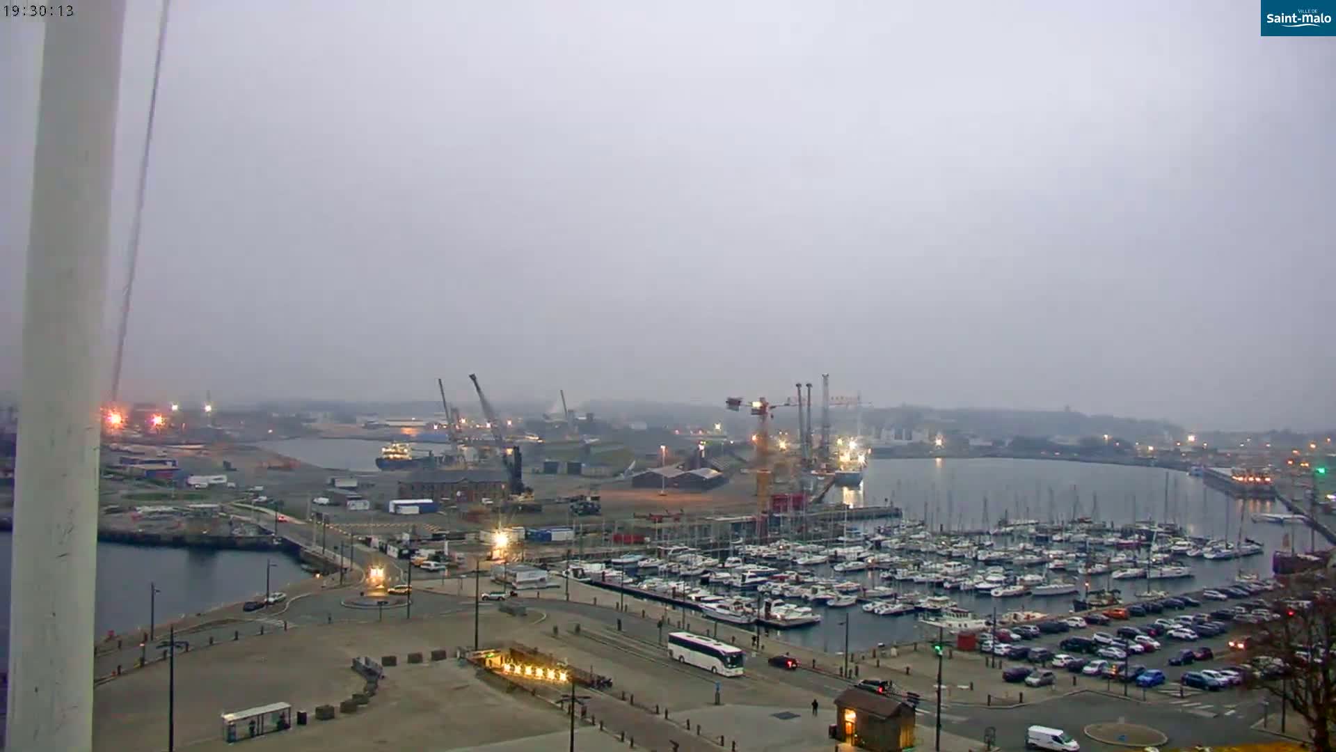 A foggy view of a harbor with numerous boats docked, industrial areas with cranes, and a parking lot with cars.