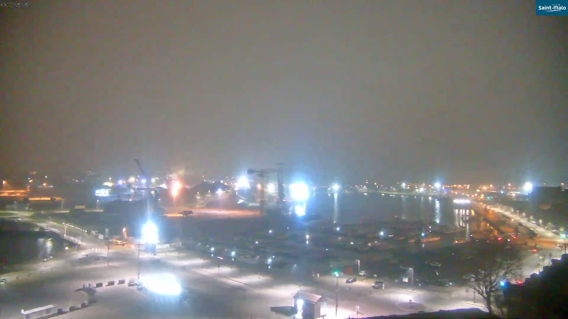 A hazy, nighttime view of a harbor with illuminated buildings, vehicles, and a large crane.