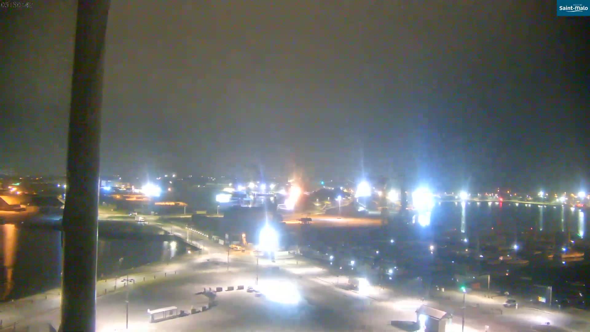 A nighttime view of a harbor and waterfront area, with lights illuminating buildings, boats, and a mostly empty parking lot, under hazy or foggy conditions.