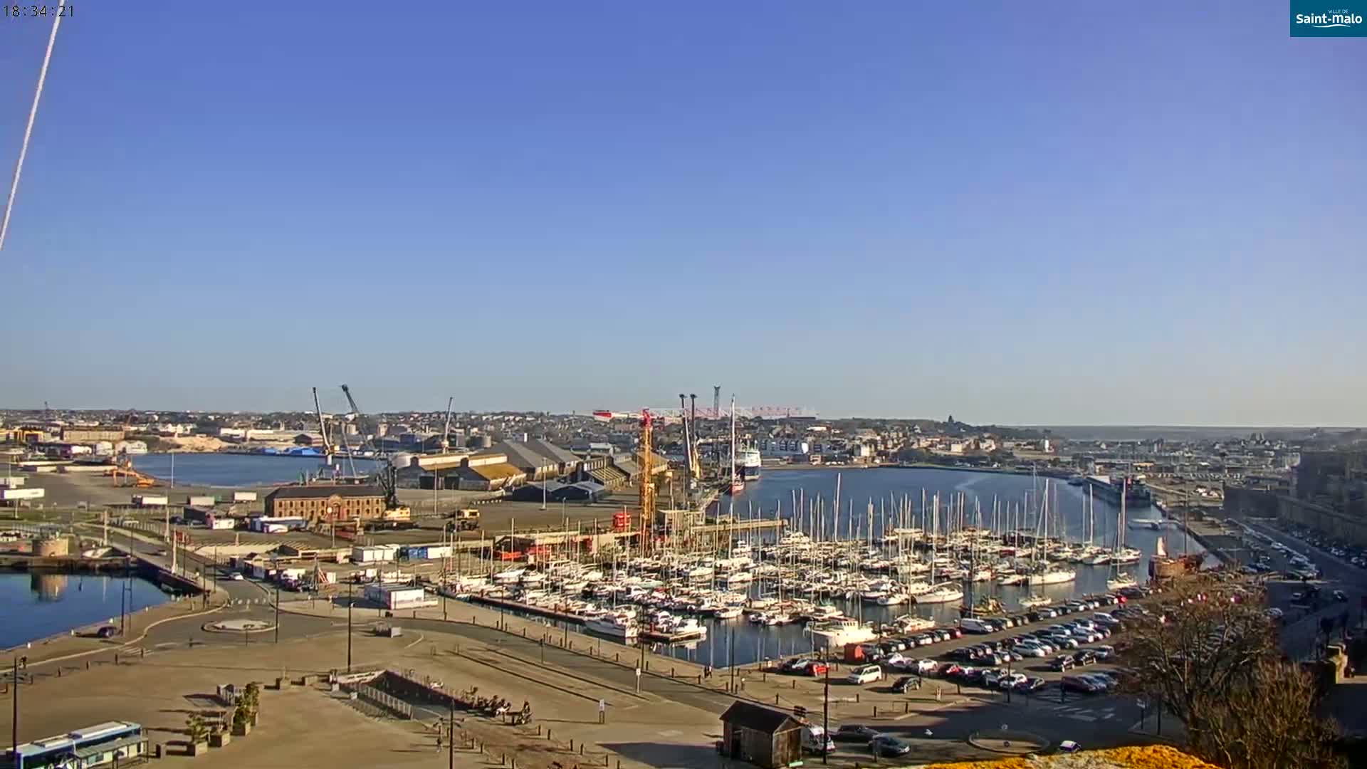 A marina filled with sailboats is situated next to a city port under a clear blue sky.