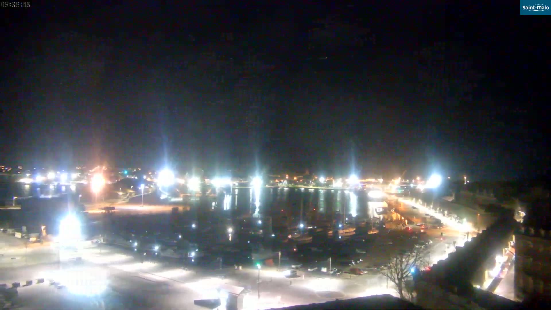 A nighttime aerial view of a harbor filled with boats, surrounded by brightly lit buildings and parking lots under a clear, dark sky.
