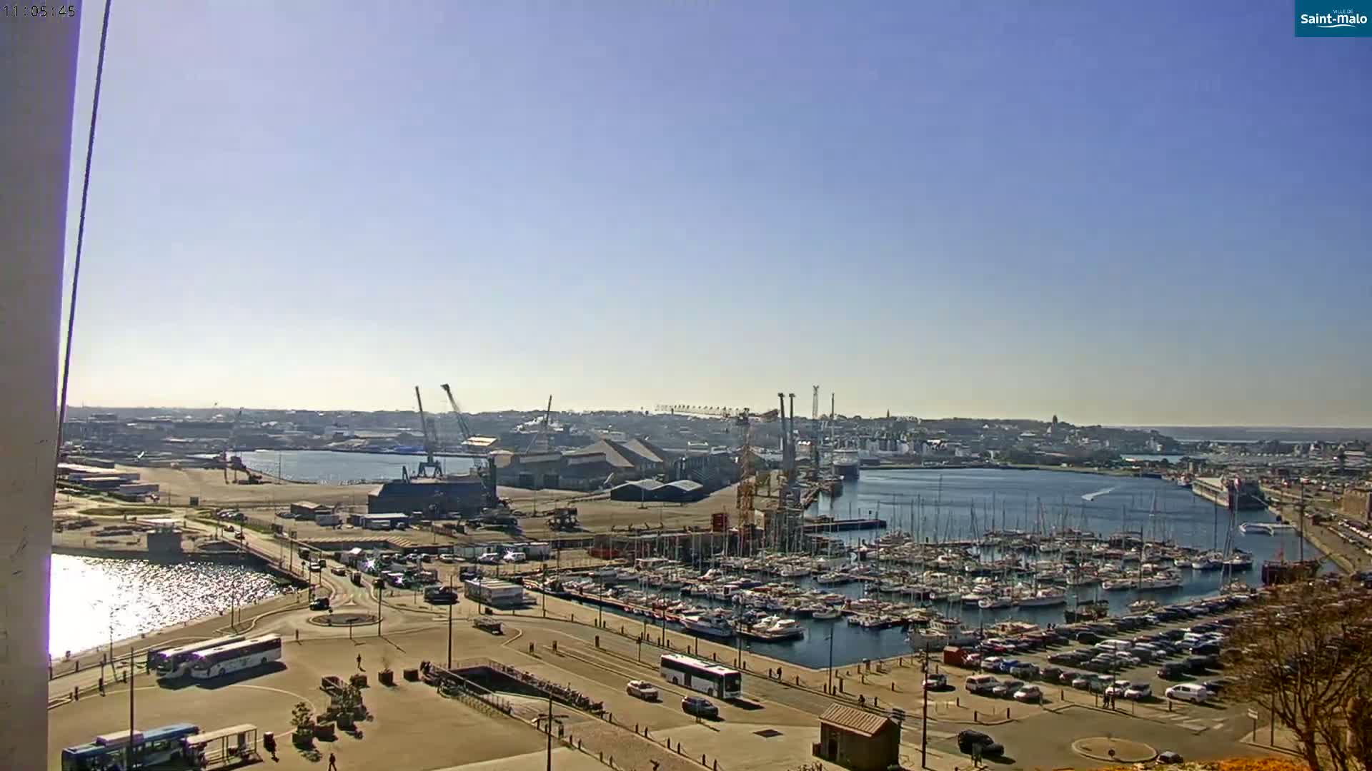 A sunny day overlooks a harbor filled with numerous boats and yachts, adjacent to a large industrial area with cranes and buildings, and a city in the distance.