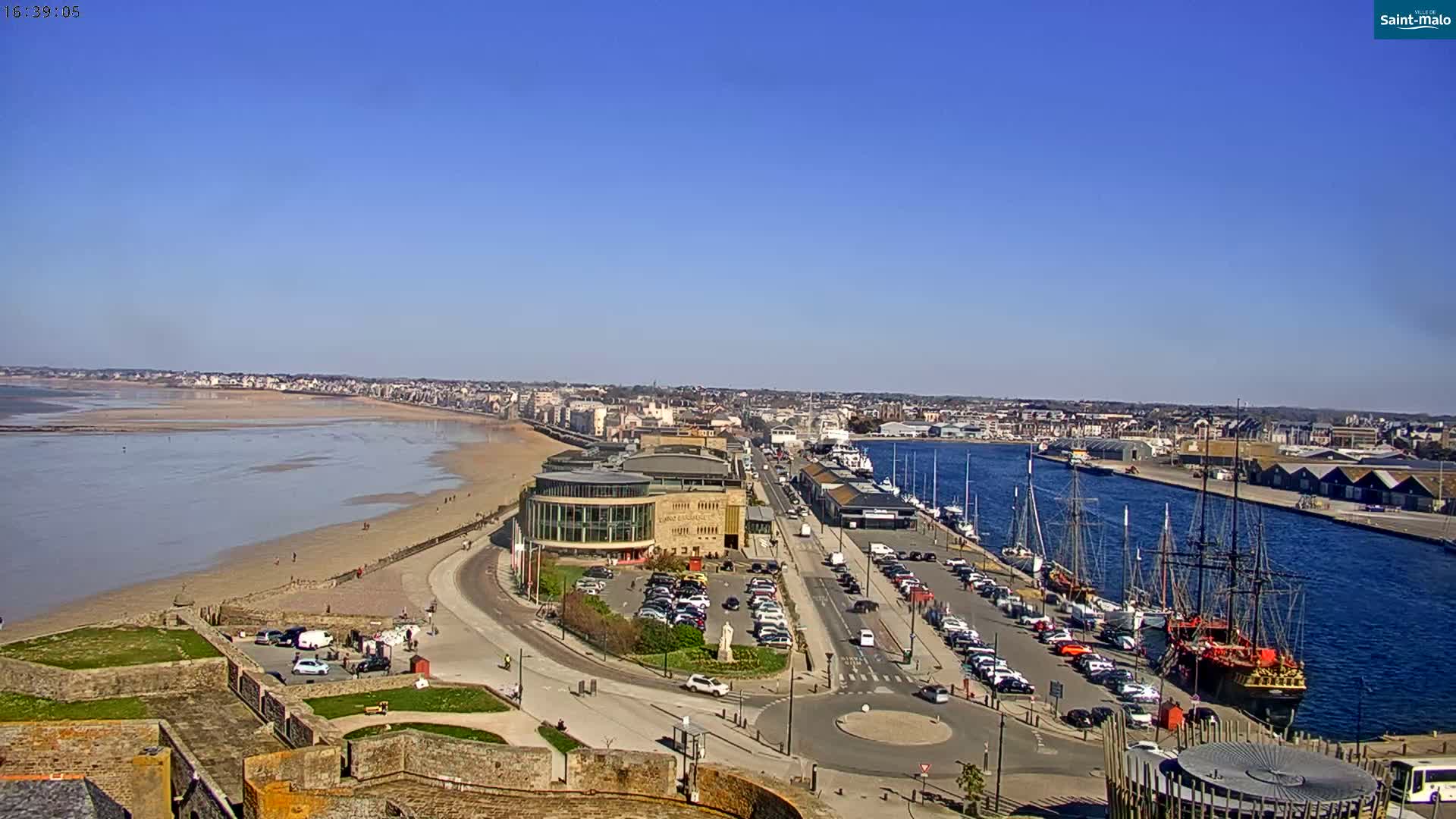 A sunny day reveals a coastal town with a sandy beach, harbor filled with boats, and a large building near the waterfront, all viewed from an elevated vantage point.