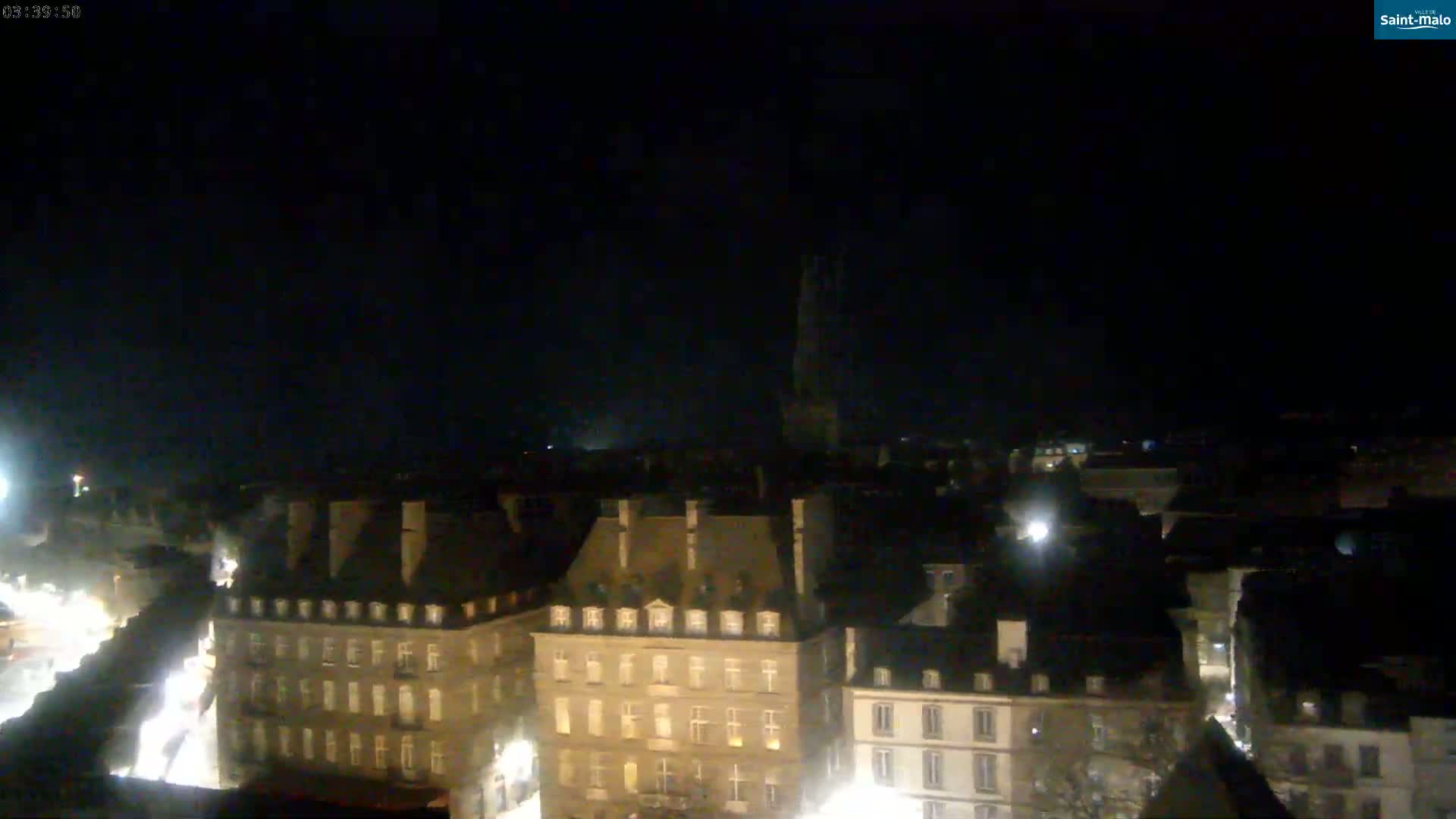 A nighttime, aerial view of a city with numerous buildings illuminated, and a tall spire visible in the distance under a dark, clear sky.