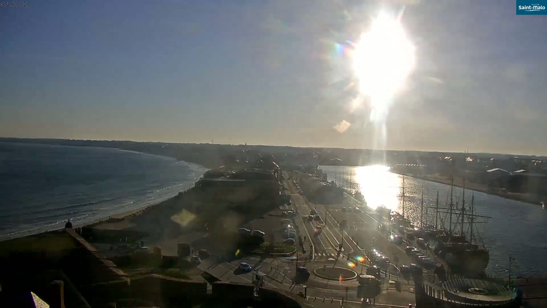 A high-angle, sunny view shows a coastal city with a harbor containing several ships, a road, and a beach with ocean waves under a mostly clear sky.