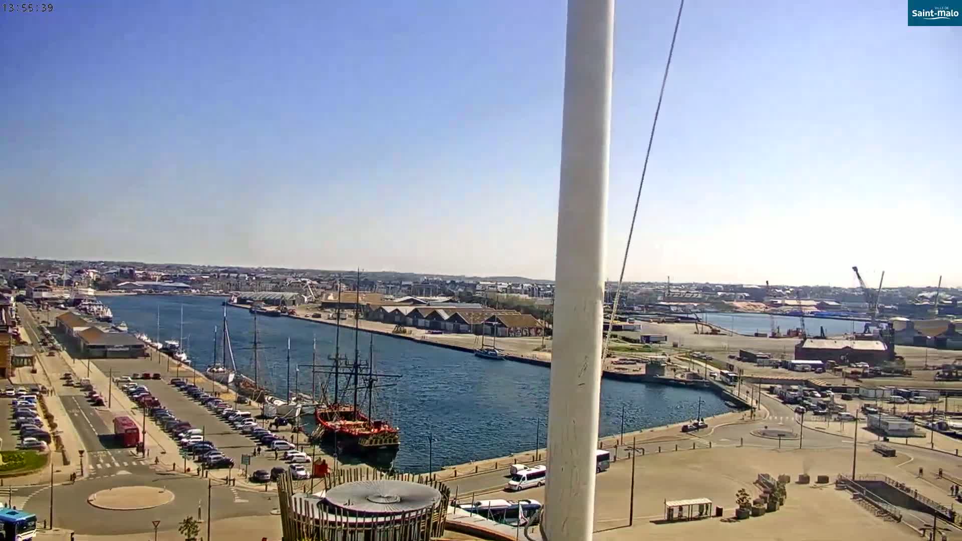 A harbor filled with docked ships and boats is viewed from an elevated perspective on a sunny day, with a city in the background and parking lots visible in the foreground.