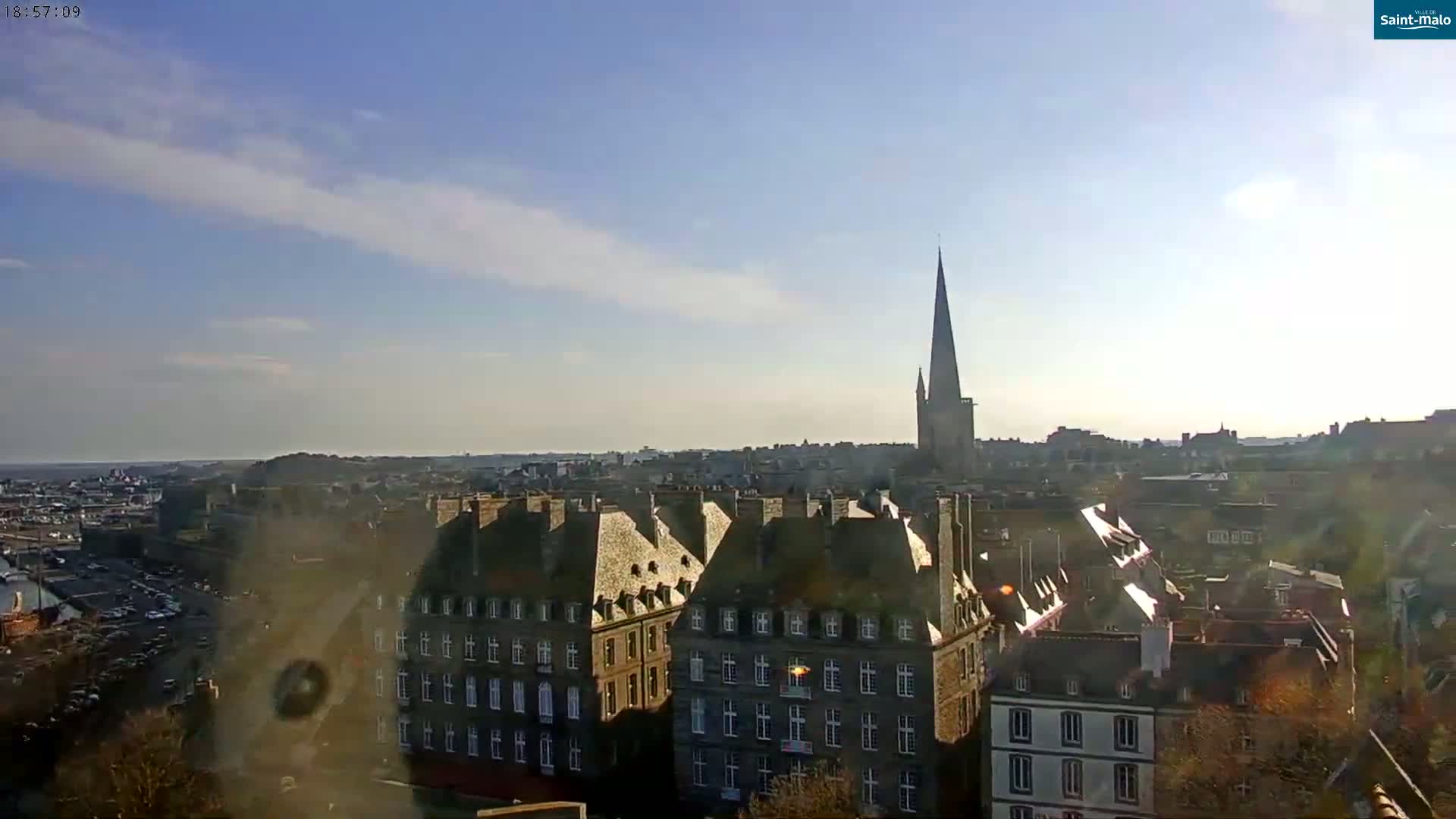 A mostly sunny cityscape with a tall church spire visible in the distance, showing many buildings with steeply pitched roofs.