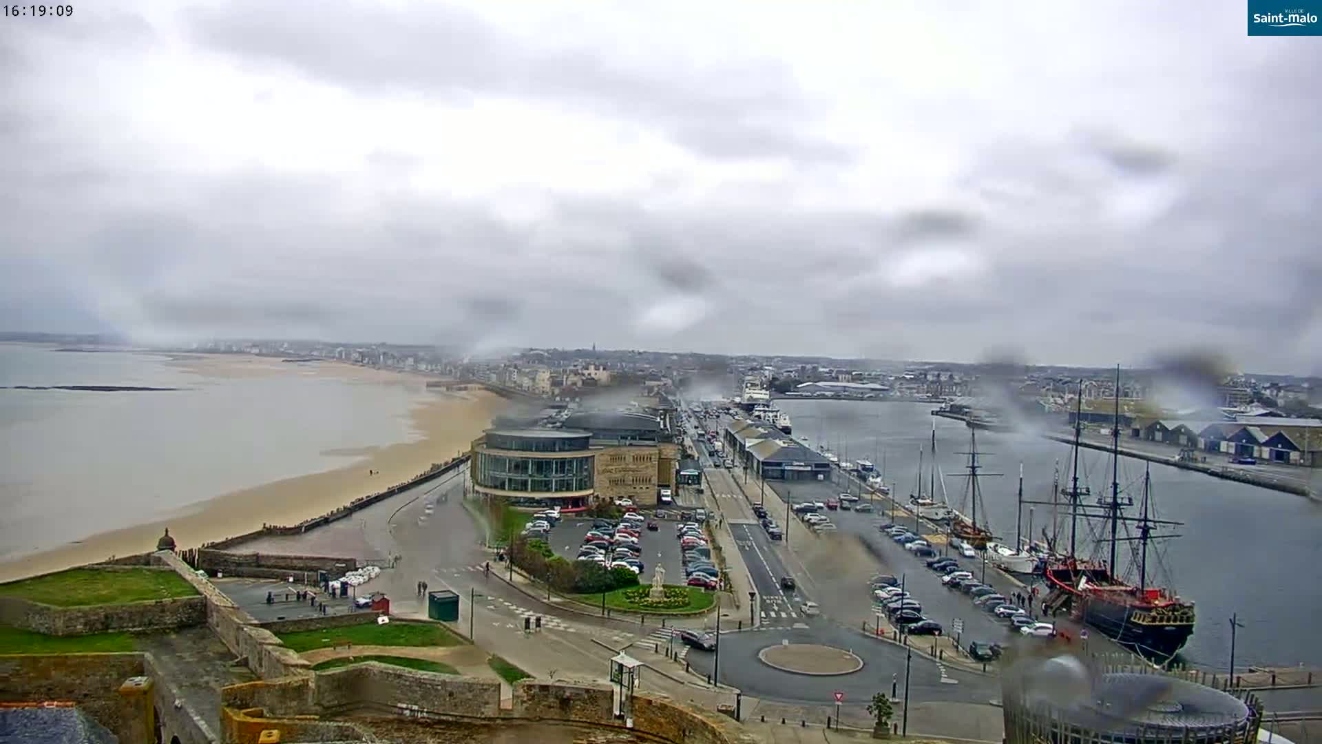 A rain-swept coastal city scene shows a harbor with several ships, a sandy beach, and a road leading to a large building, all viewed from an elevated perspective.