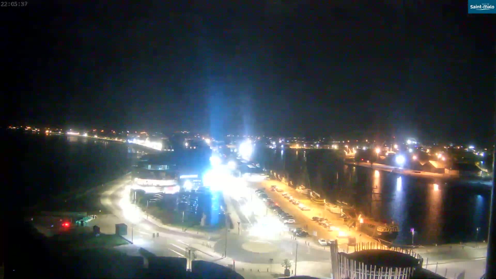 A nighttime view of a waterfront city illuminated by streetlights and building lights, with calm water reflecting the lights.