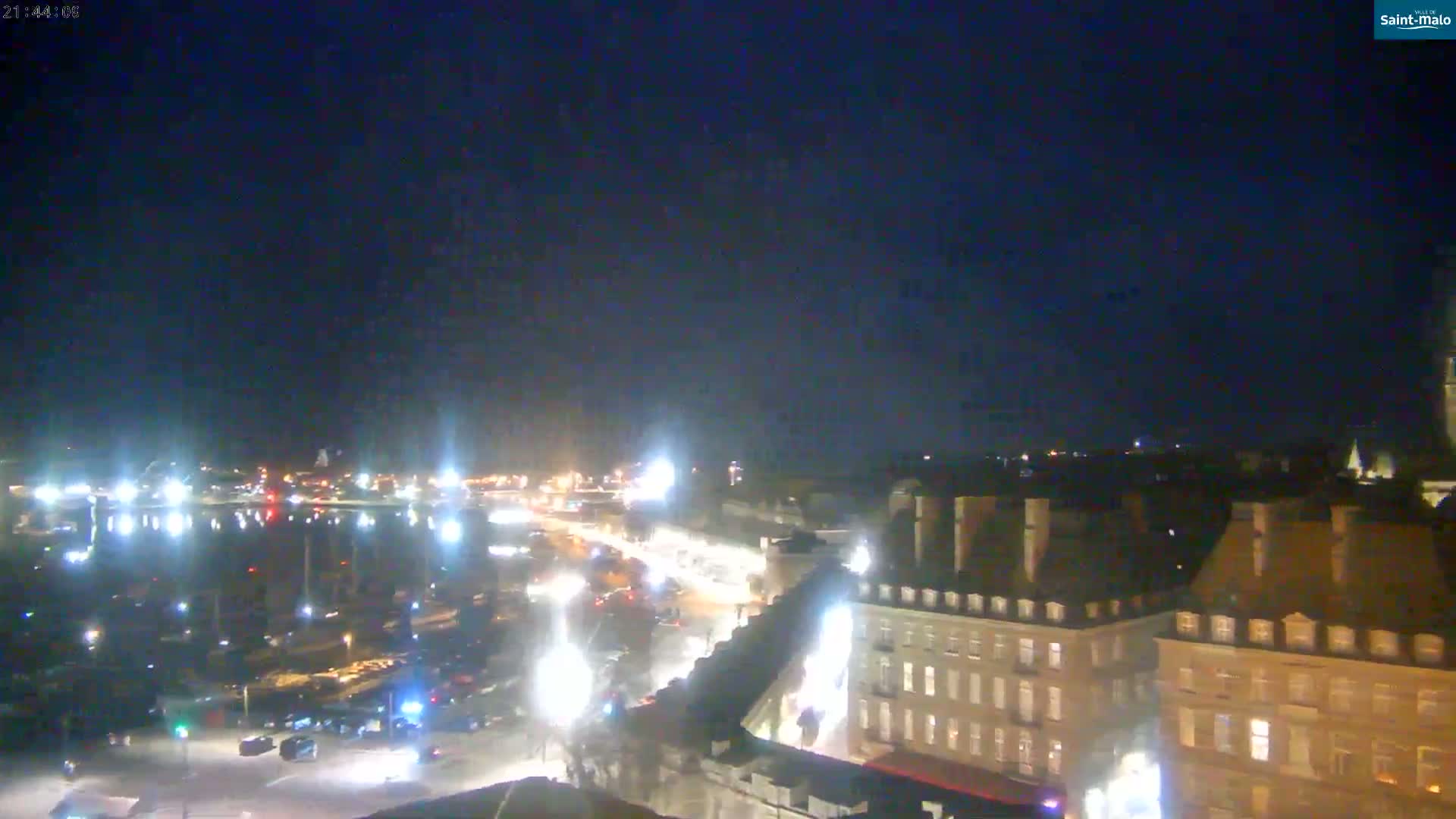 A nighttime, high-angle view shows a city harbor with illuminated buildings and vehicles along a roadway.