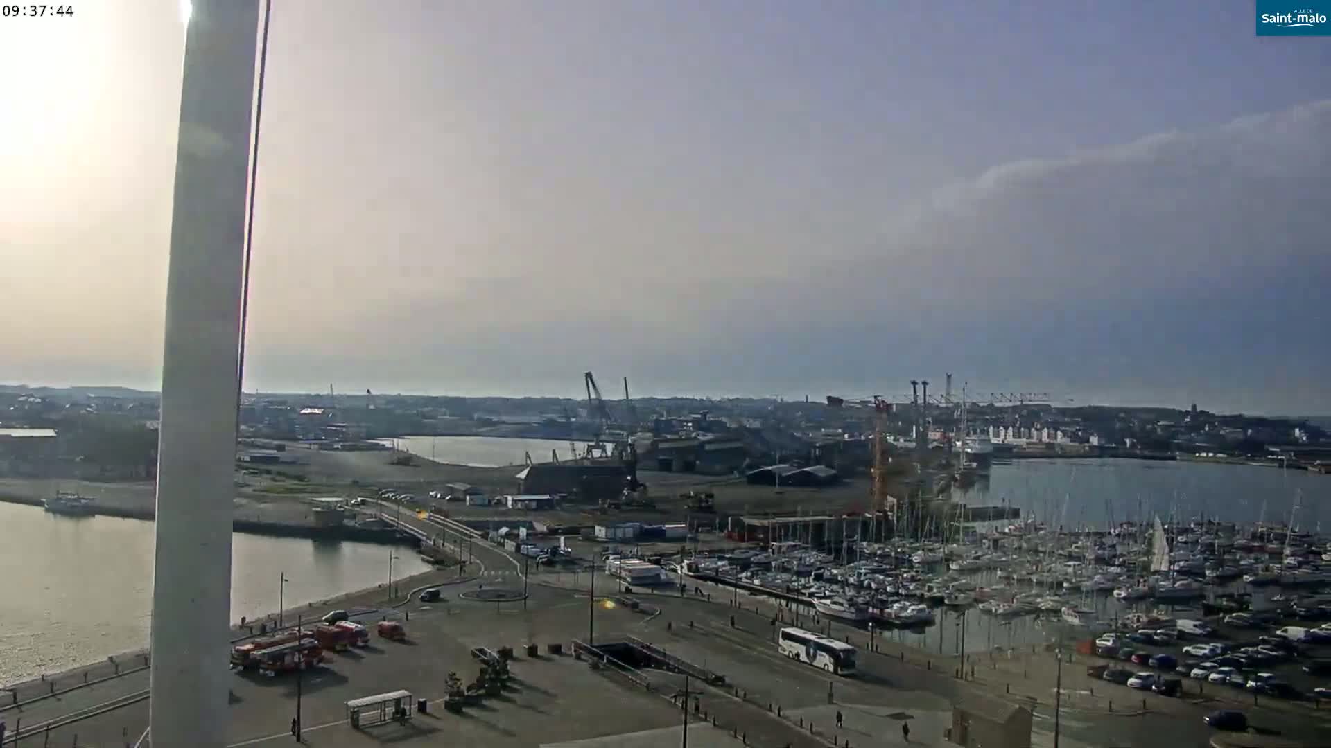 A high-angle view shows a harbor filled with numerous sailboats and yachts, industrial areas, and a city under a mostly clear, slightly hazy sky.