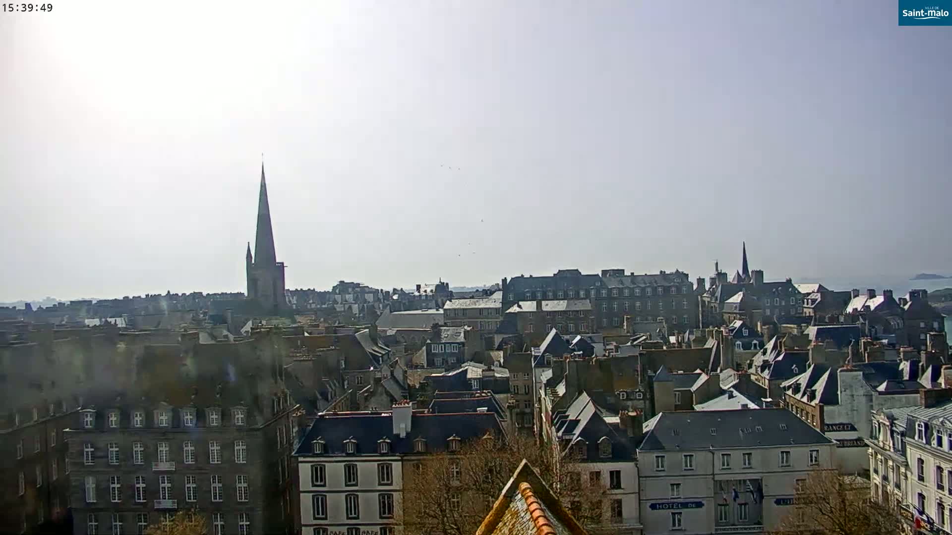 A hazy, sunny day overlooks a cityscape of many closely-packed buildings with dark gray slate roofs, punctuated by a tall, pointed church spire.